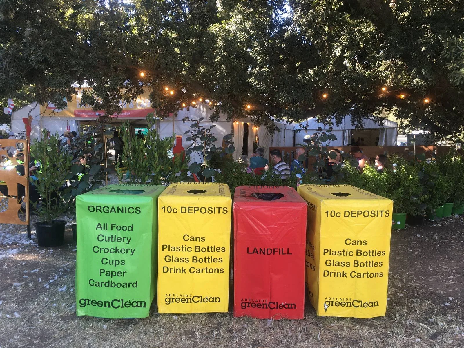 Recycling bins in green, yellow, and red labeled for organic food waste, deposits, and landfill respectively, with a background of trees, string lights, and people sitting at an outdoor event under a tent.