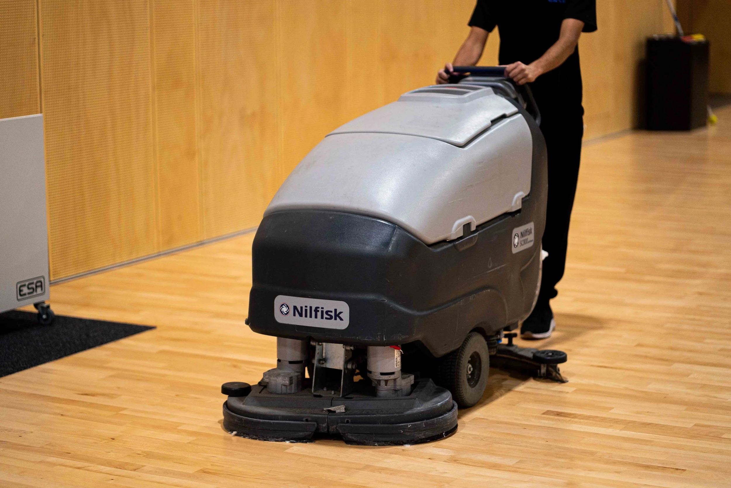 A person operating a Nilfisk floor cleaning machine in a room with wooden floors and orange wood-paneled walls.