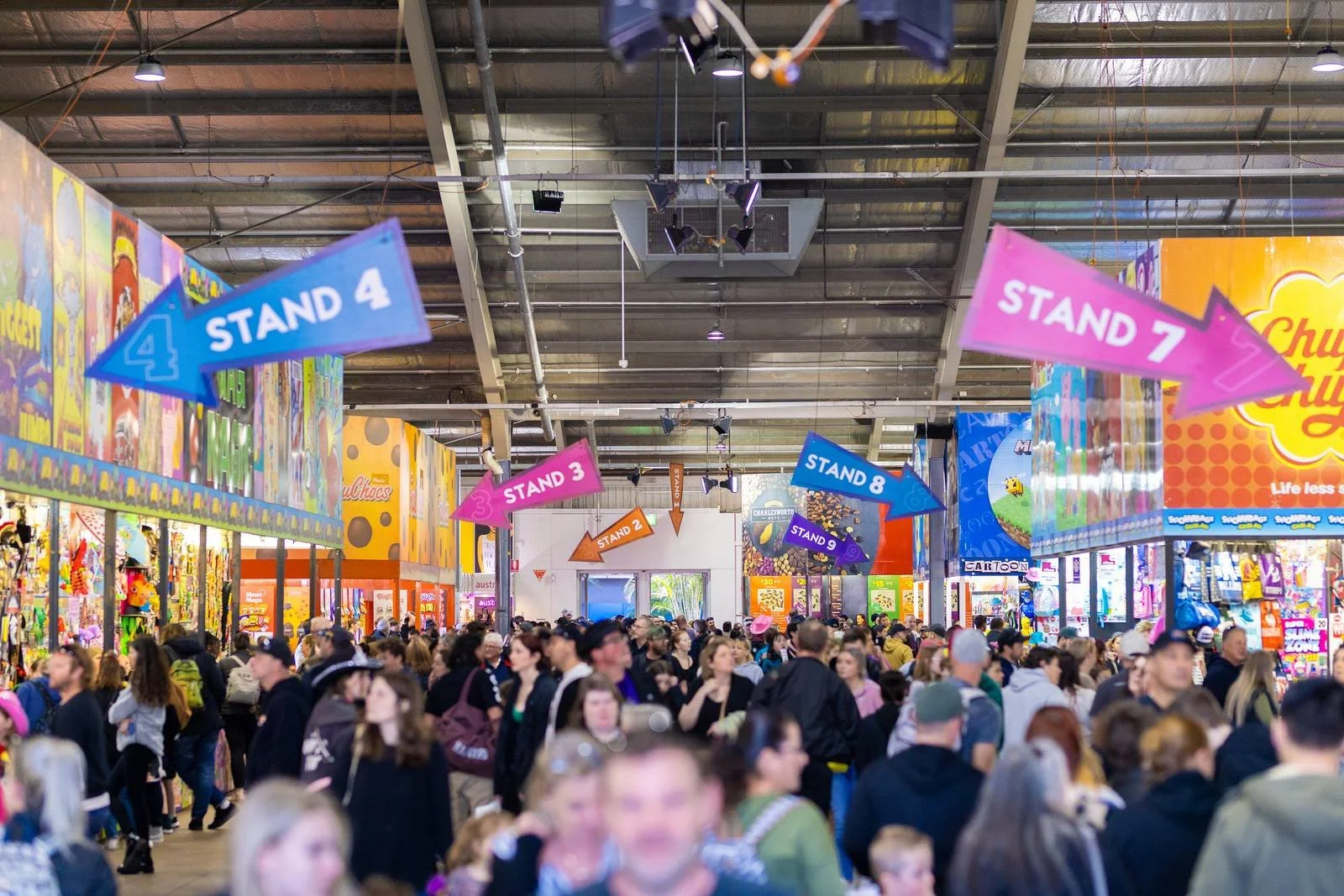 Crowd of people at a convention or event with colorful booth displays and hanging directional signs indicating different stands numbered 1 to 9.