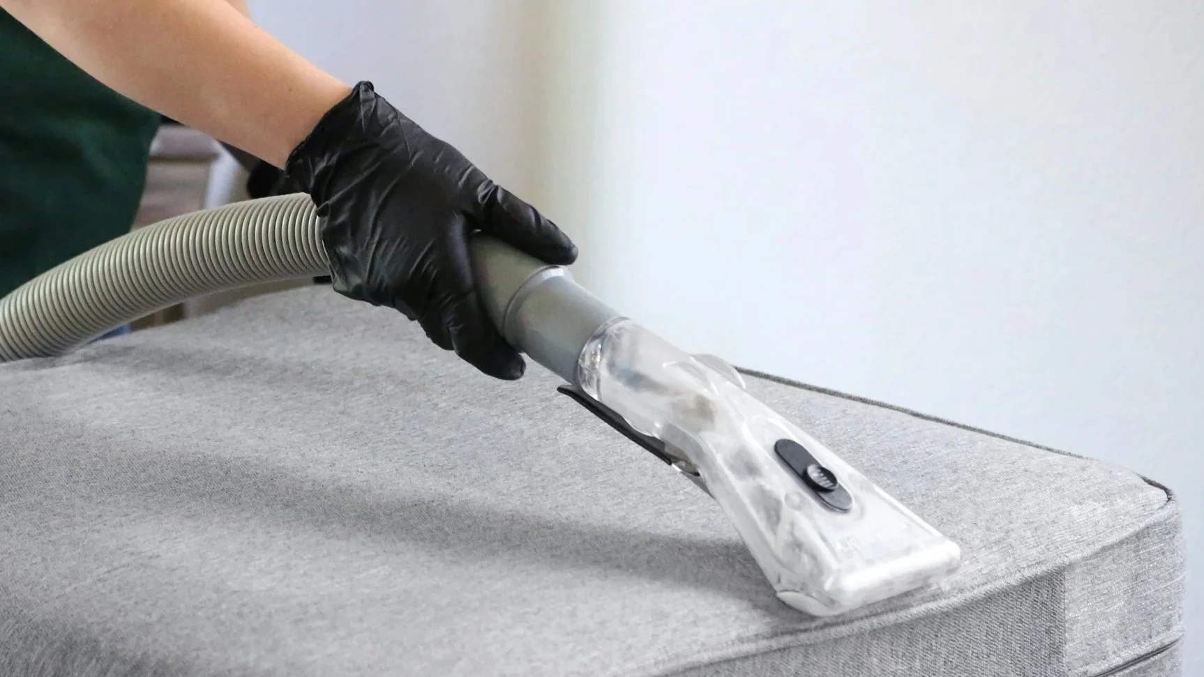 Person wearing black gloves vacuuming a light gray fabric surface with a handheld vacuum cleaner.