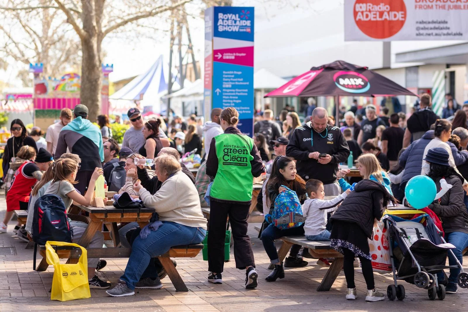 A crowded outdoor community event with people sitting at wooden tables, children, families, and volunteers. Tents, an amusement ride, and signs for the Royal Adelaide Show and radio station are visible.