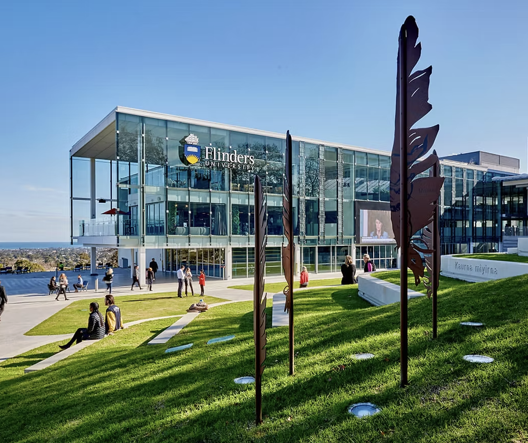 Exterior of Flinders University with glass building, outdoor seating, green lawn, metal feather sculptures, and people walking and sitting outside on a sunny day.