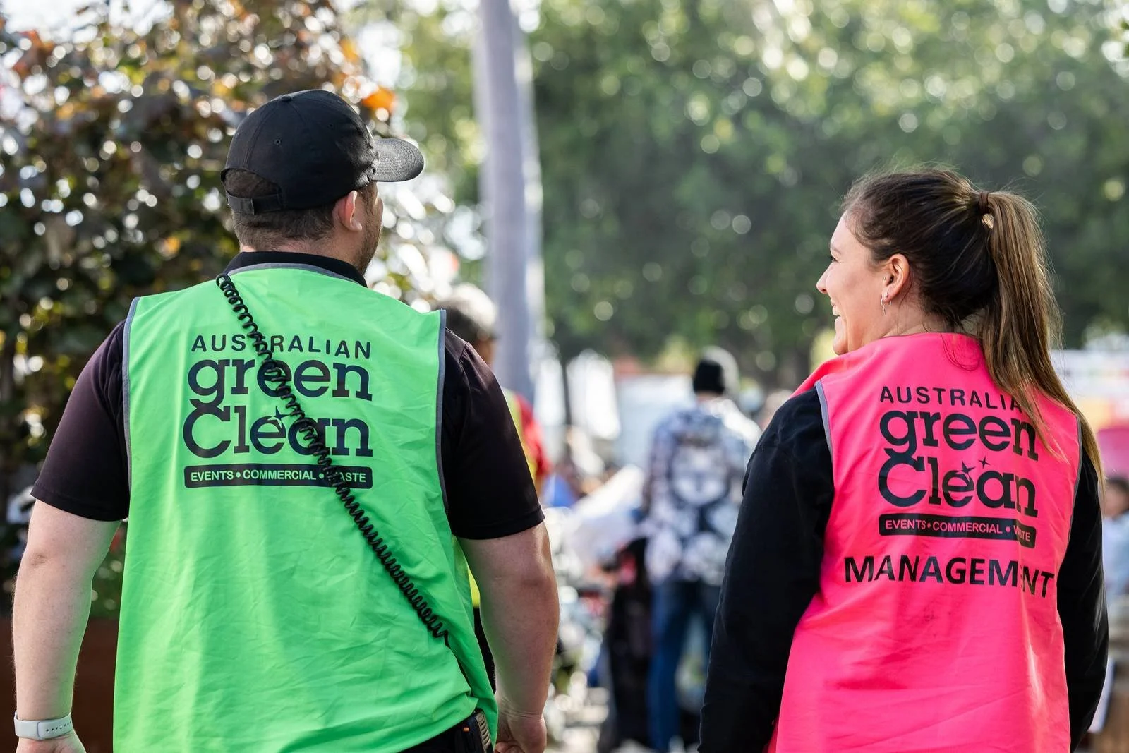 Two people wearing high-visibility jackets with 'Australian Green Clean' printed on the back, talking outdoors during a daytime event. The person on the left is a man with a black cap and a green jacket, and the person on the right is a woman with a ponytail and a pink jacket.