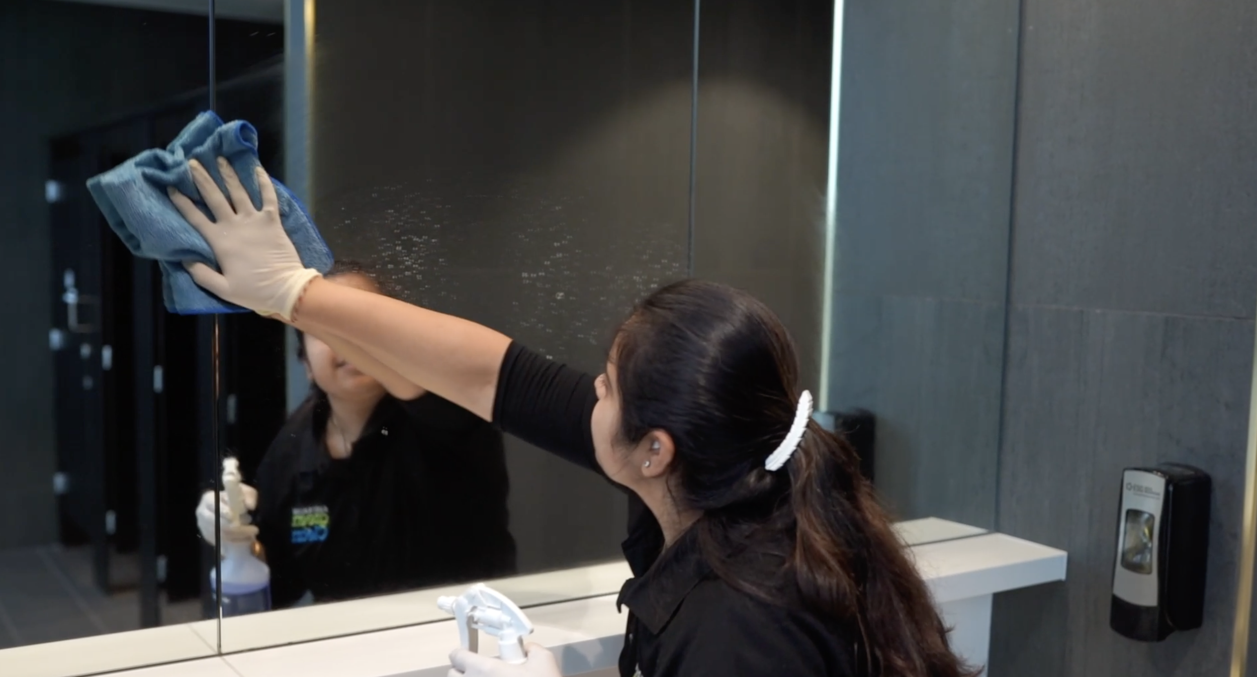 A woman cleaning a mirror in a restroom using a blue cloth, with her reflection visible in the mirror.