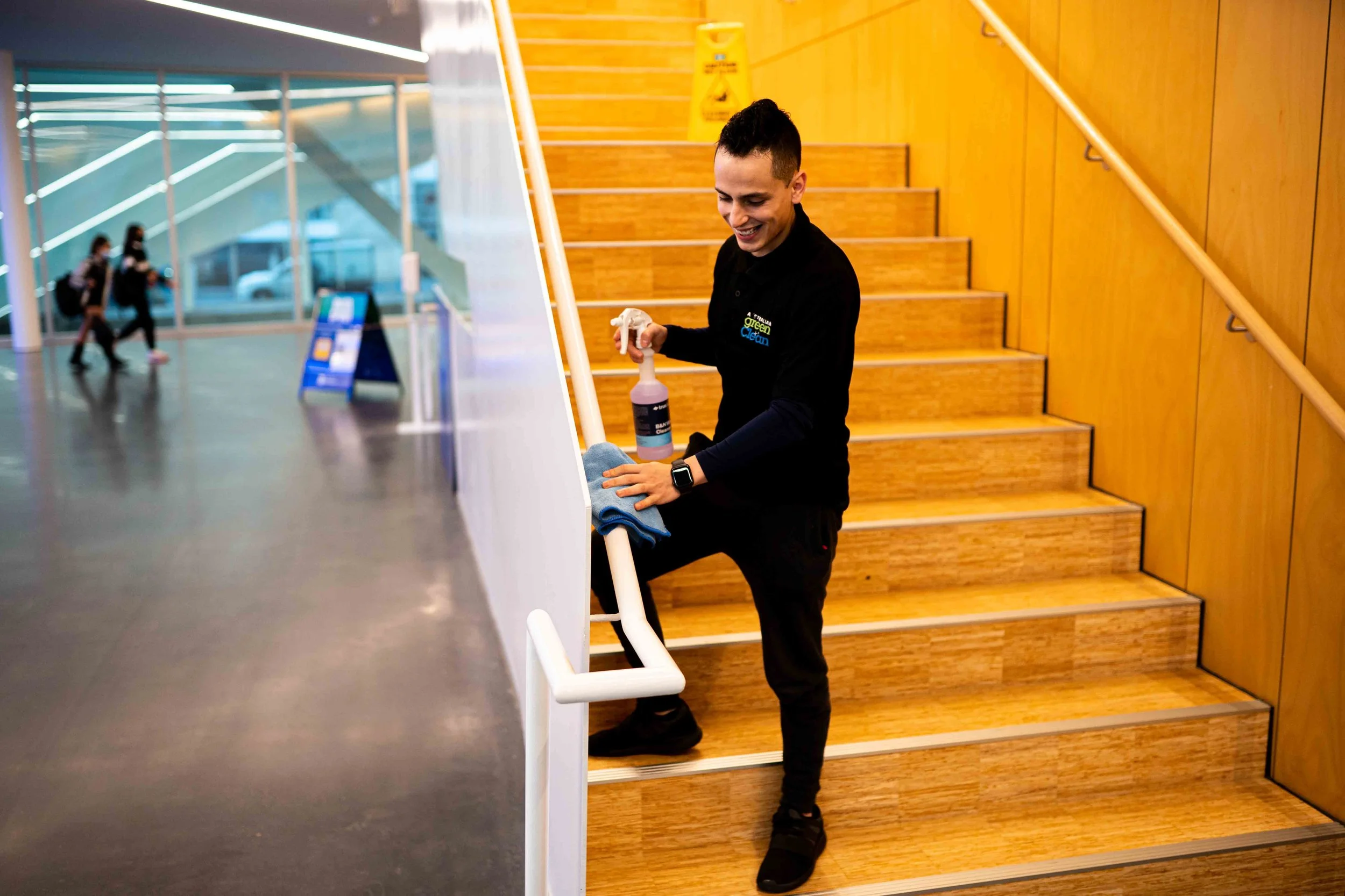 A person with short dark hair is cleaning a staircase in an indoor public space. They are smiling, wearing black clothing, and holding a spray bottle and a cloth. The staircase is made of wood, and the background includes a wooden wall and a glass window with two people walking past.