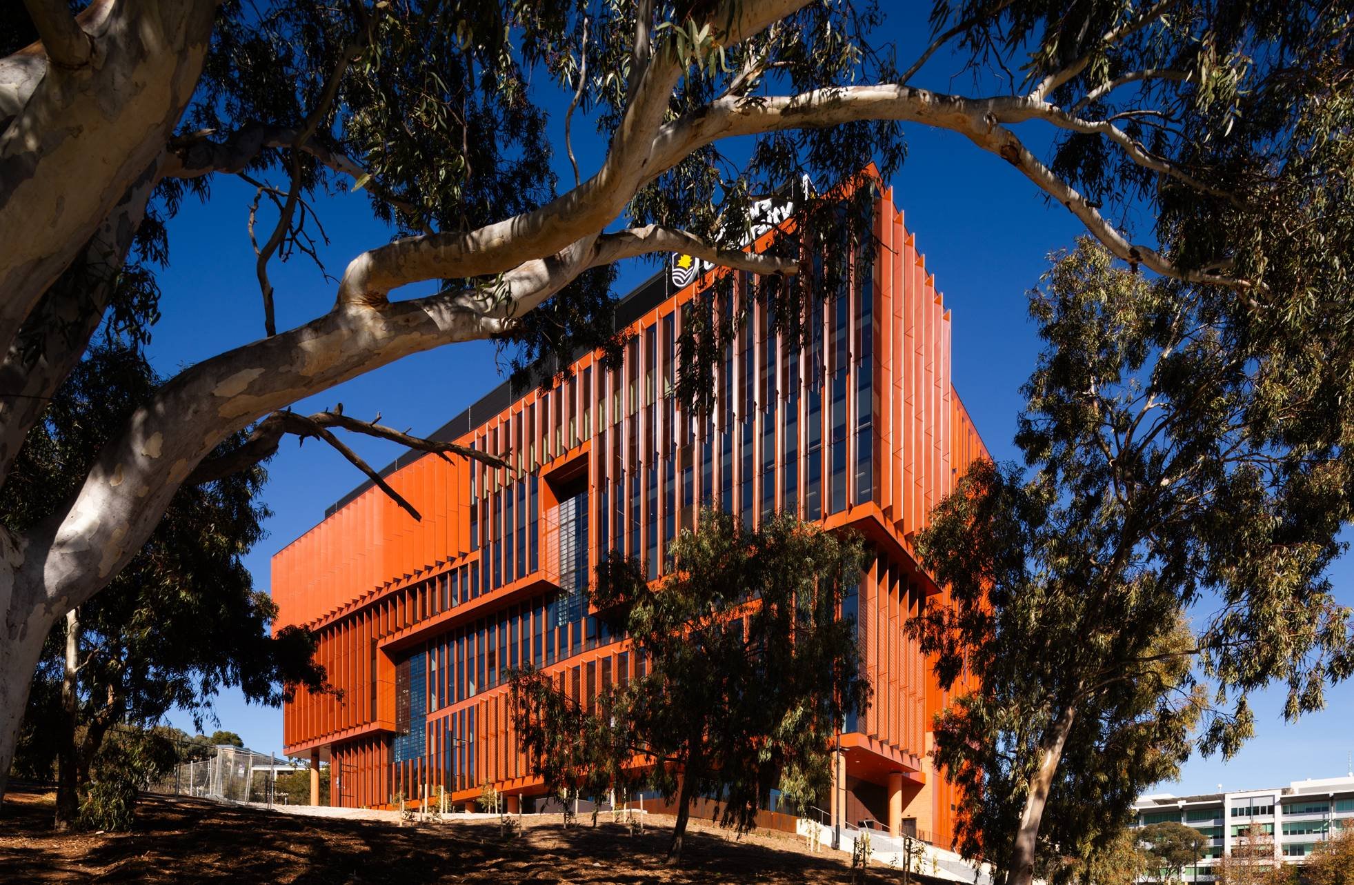 A modern orange and glass office building surrounded by trees with green leaves, set against a clear blue sky.