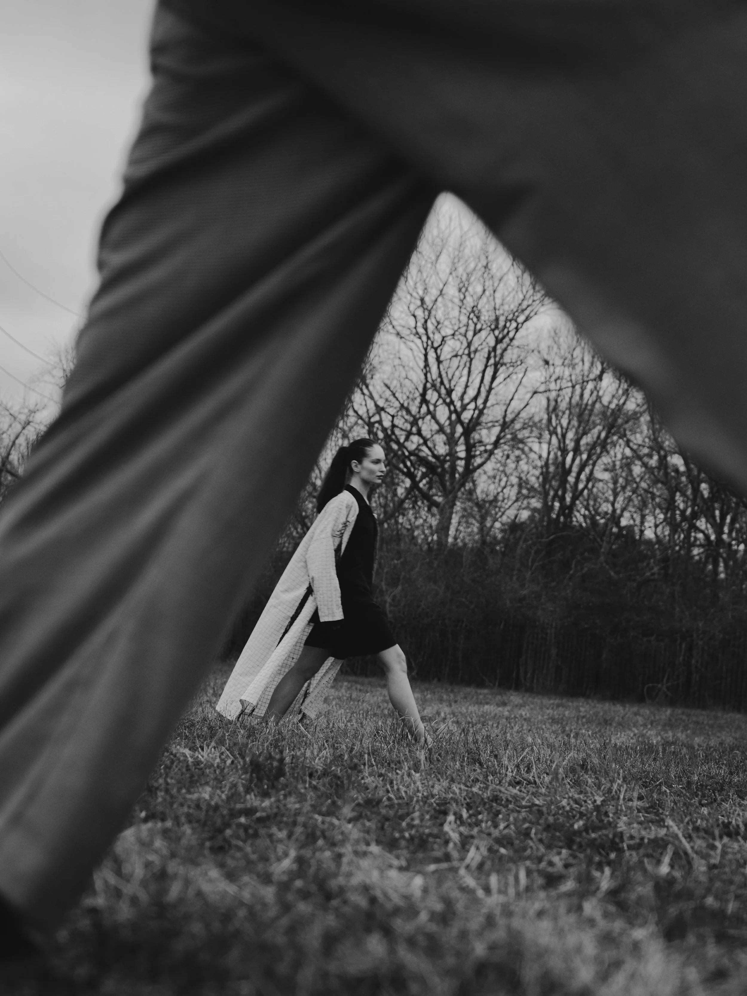 Black and white photograph of a woman walking outdoors in a park with leafless trees in the background. The image is taken from a low angle, partially obscured by a large dark object in the foreground.