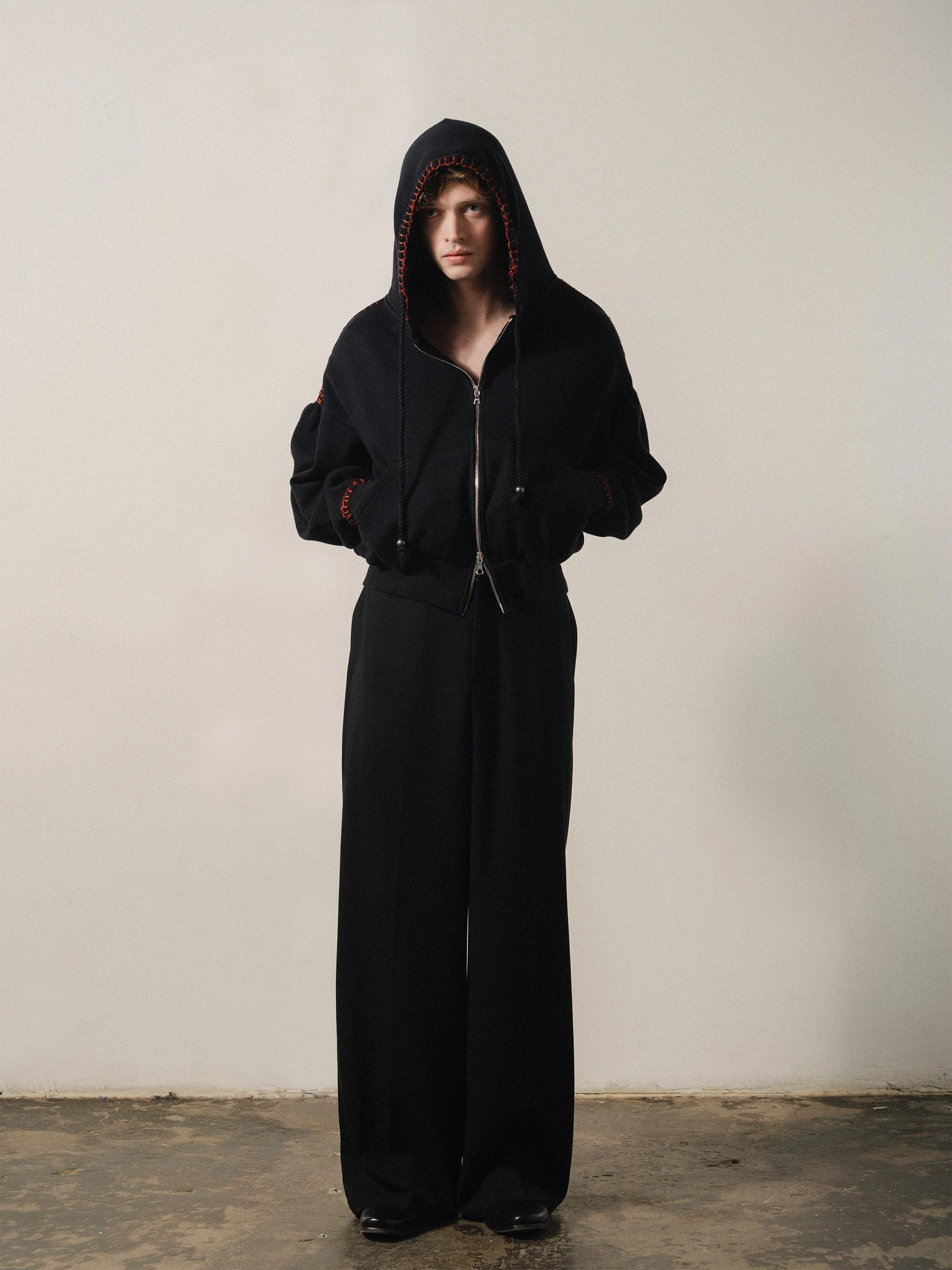 A person standing against a plain wall, dressed in a black hoodie with red stitching and loose black pants. The person has long sleeves, hands in the hoodie pockets, and wears black shoes. The hoodie has drawstrings, and the person's face is visible under the hood.