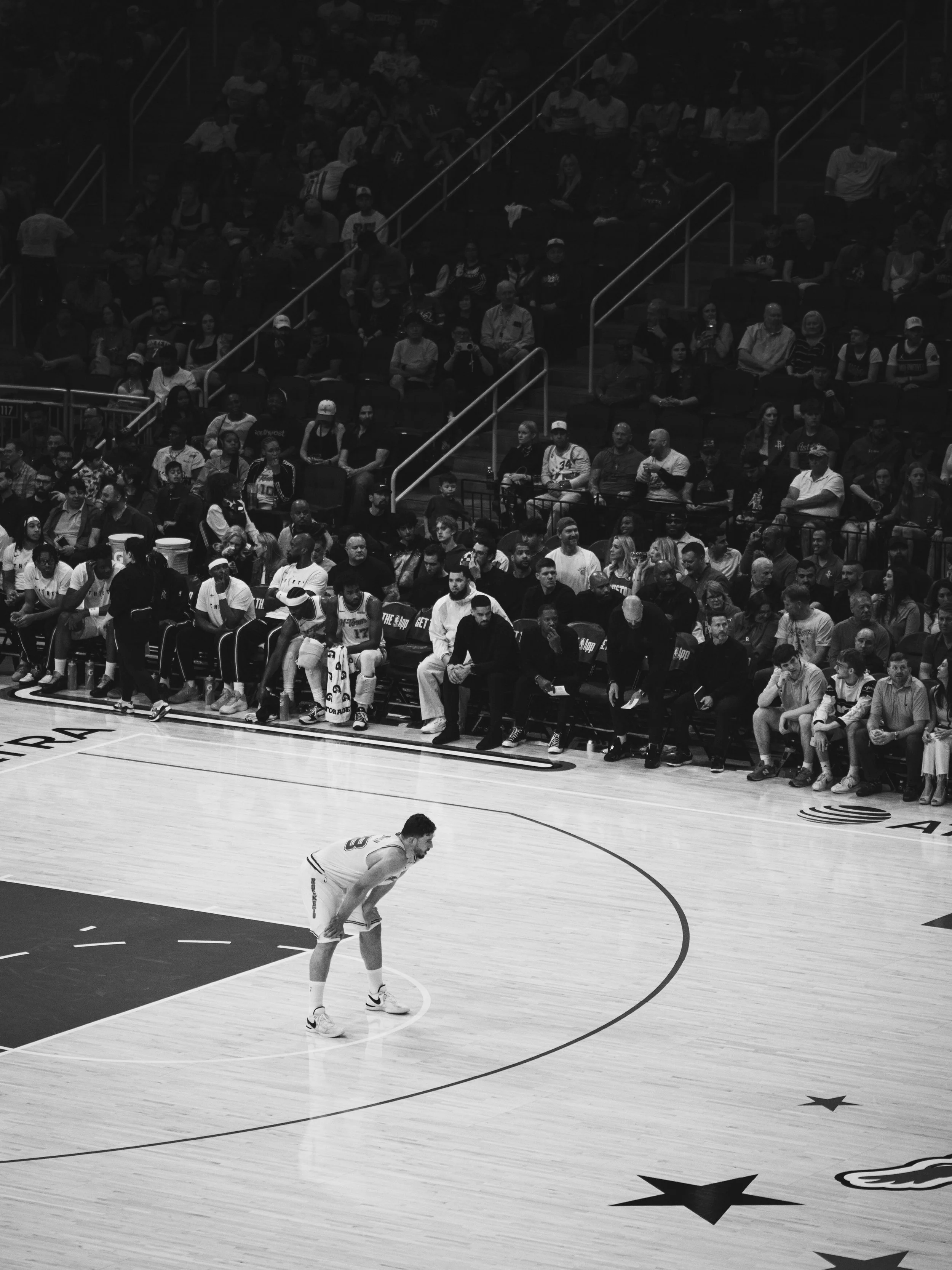 A basketball player in a white uniform stands on the court, preparing for a free throw, while spectators watch from the stands.