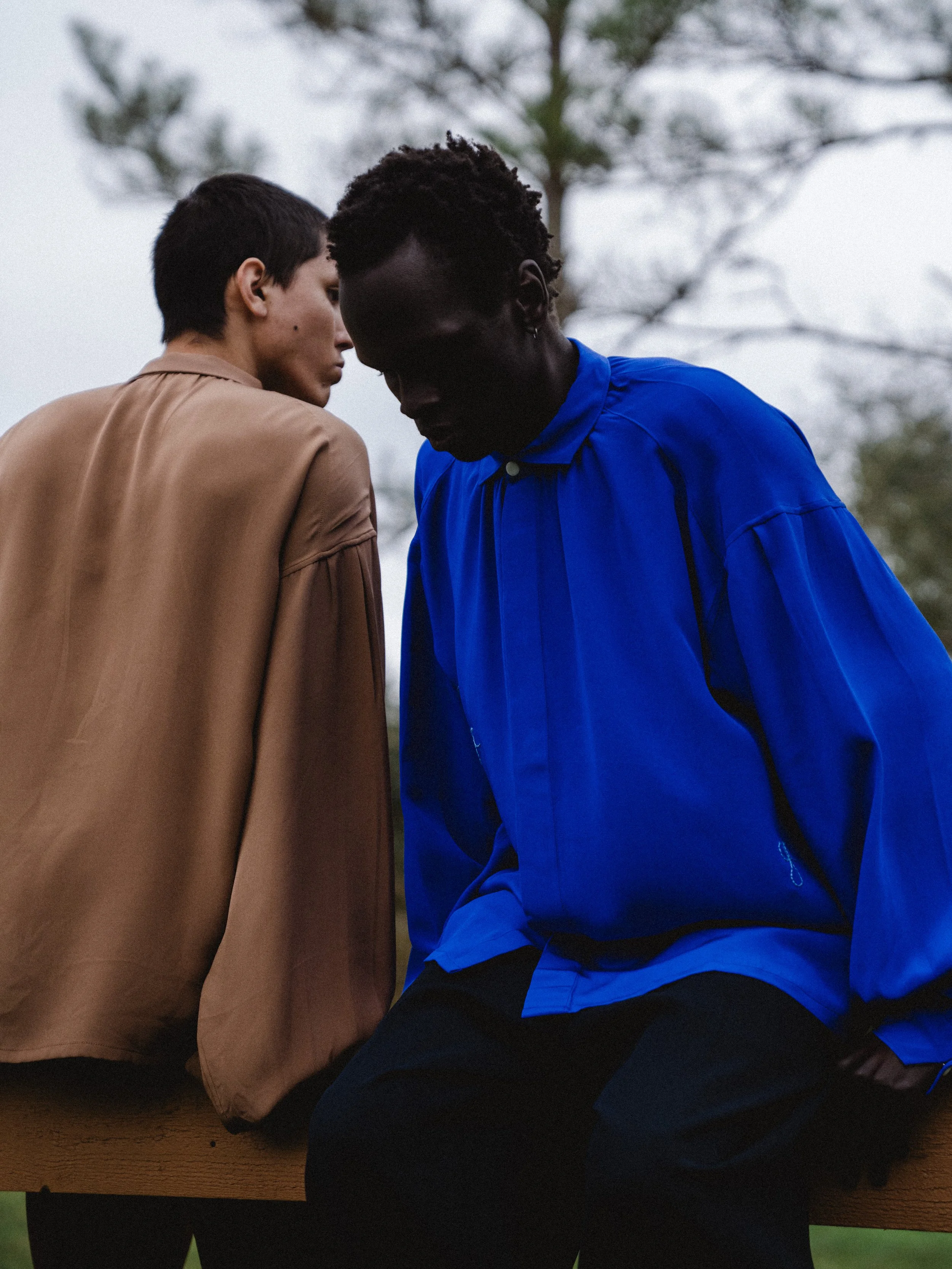 Two individuals sitting close together, one wearing a tan jacket and the other wearing a bright blue jacket, with blurred trees in the background.