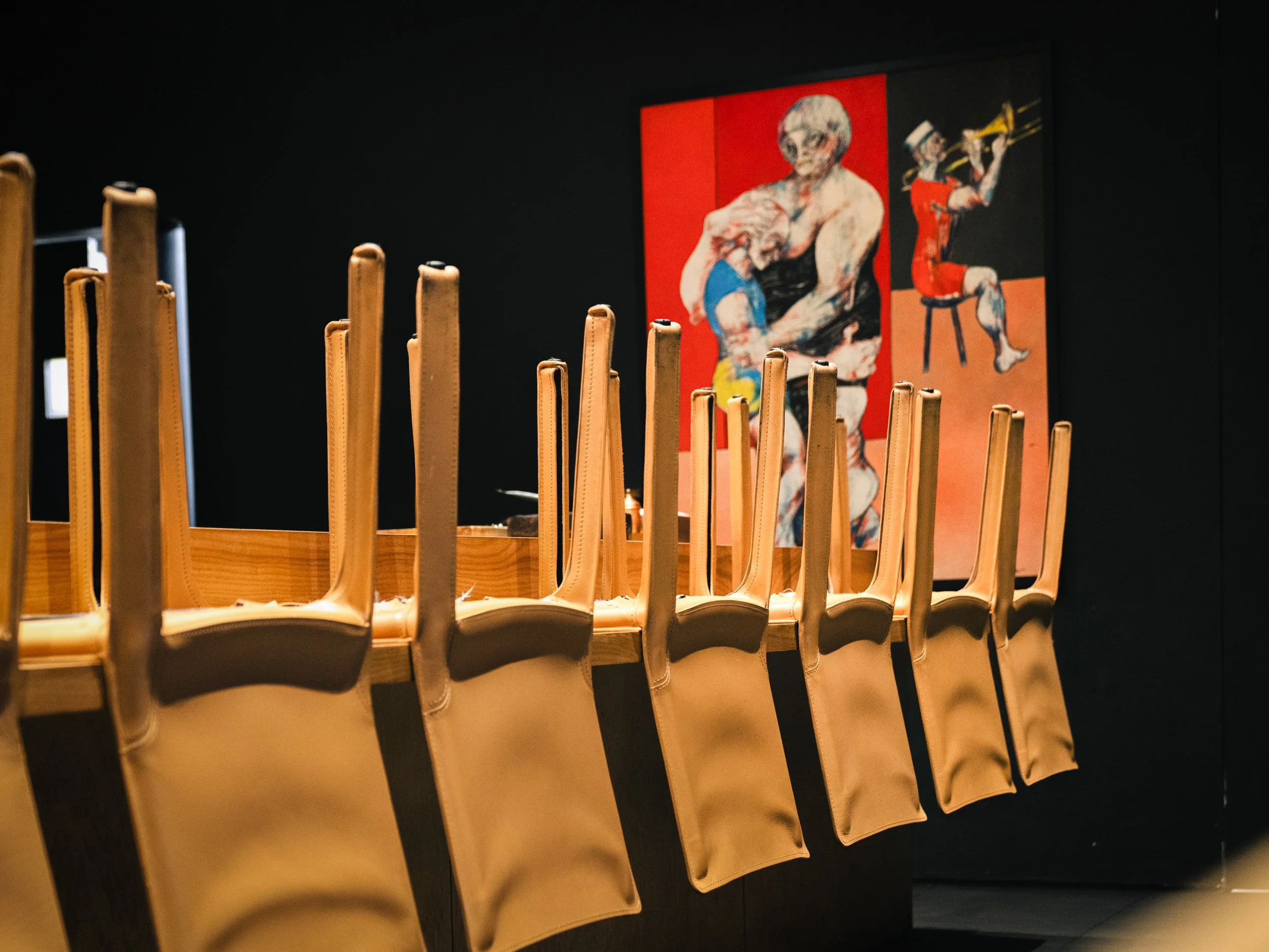 Row of empty beige chairs with black seats in a gallery or museum, with colorful artwork on the wall in the background.