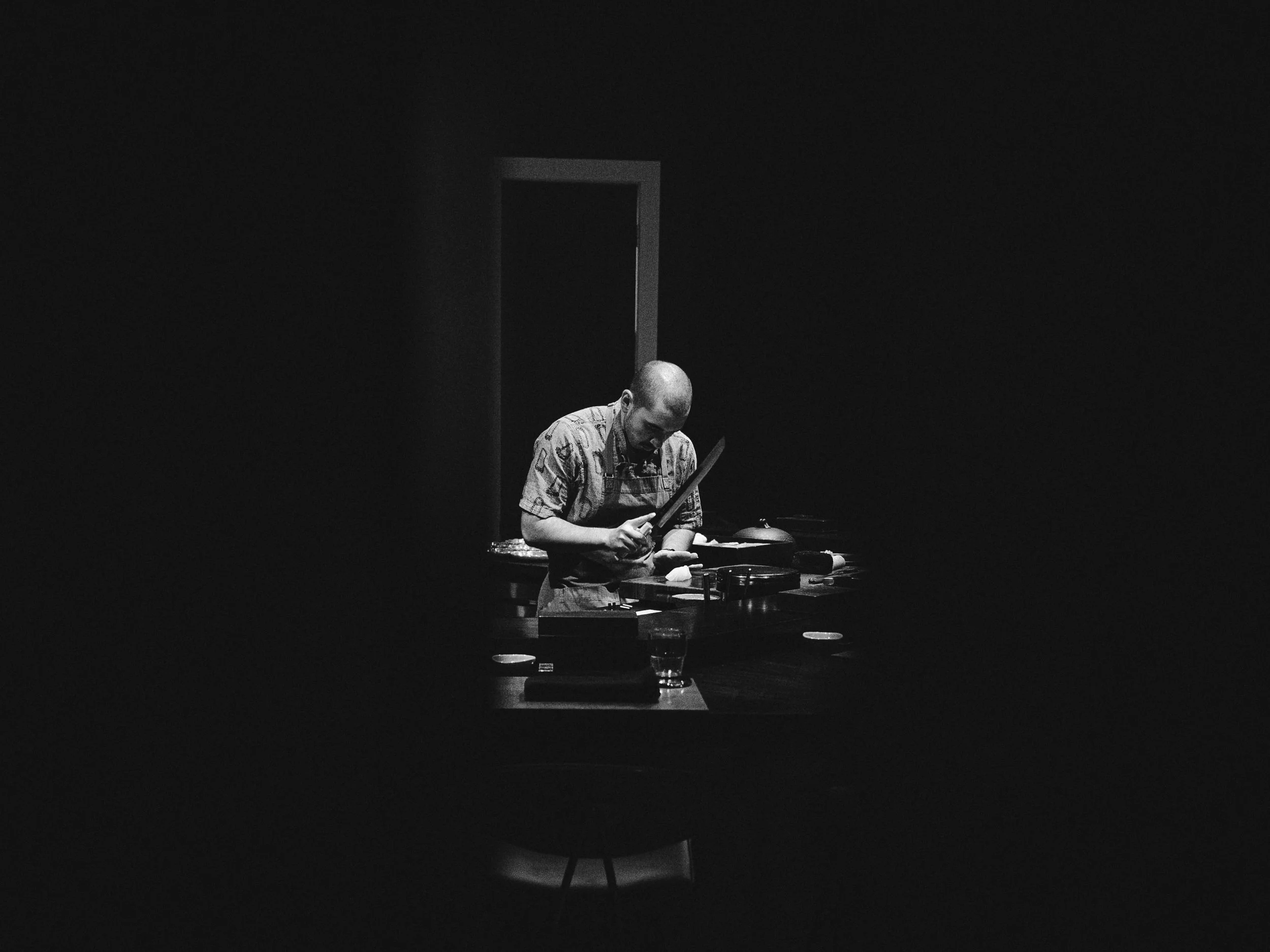 A man wearing an apron standing at a table in a dark room, illuminated by a single light, with a glass of water and some items on the table.