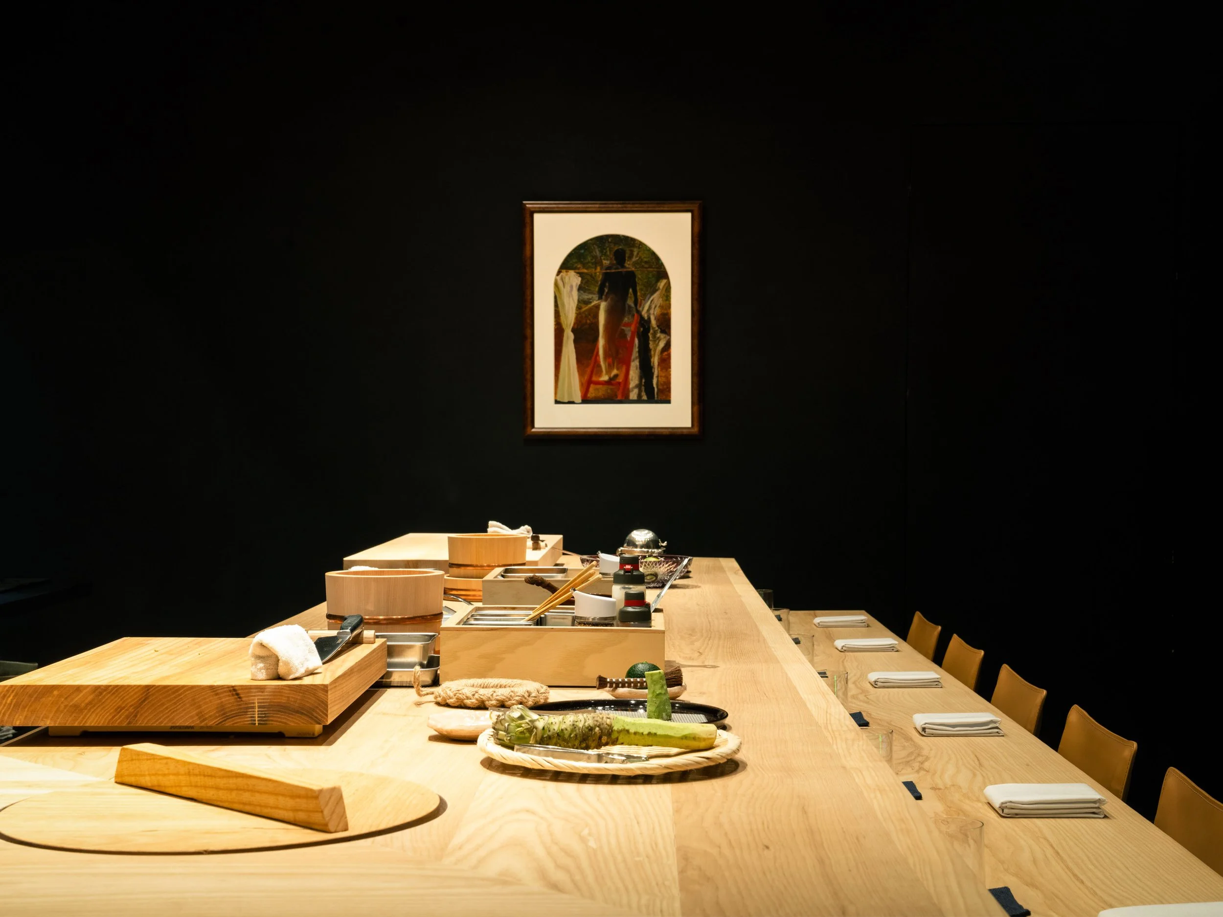 A wooden table set with plates and napkins, with various kitchen utensils and ingredients, in a room with dark walls and a framed artwork of a person standing outdoors.