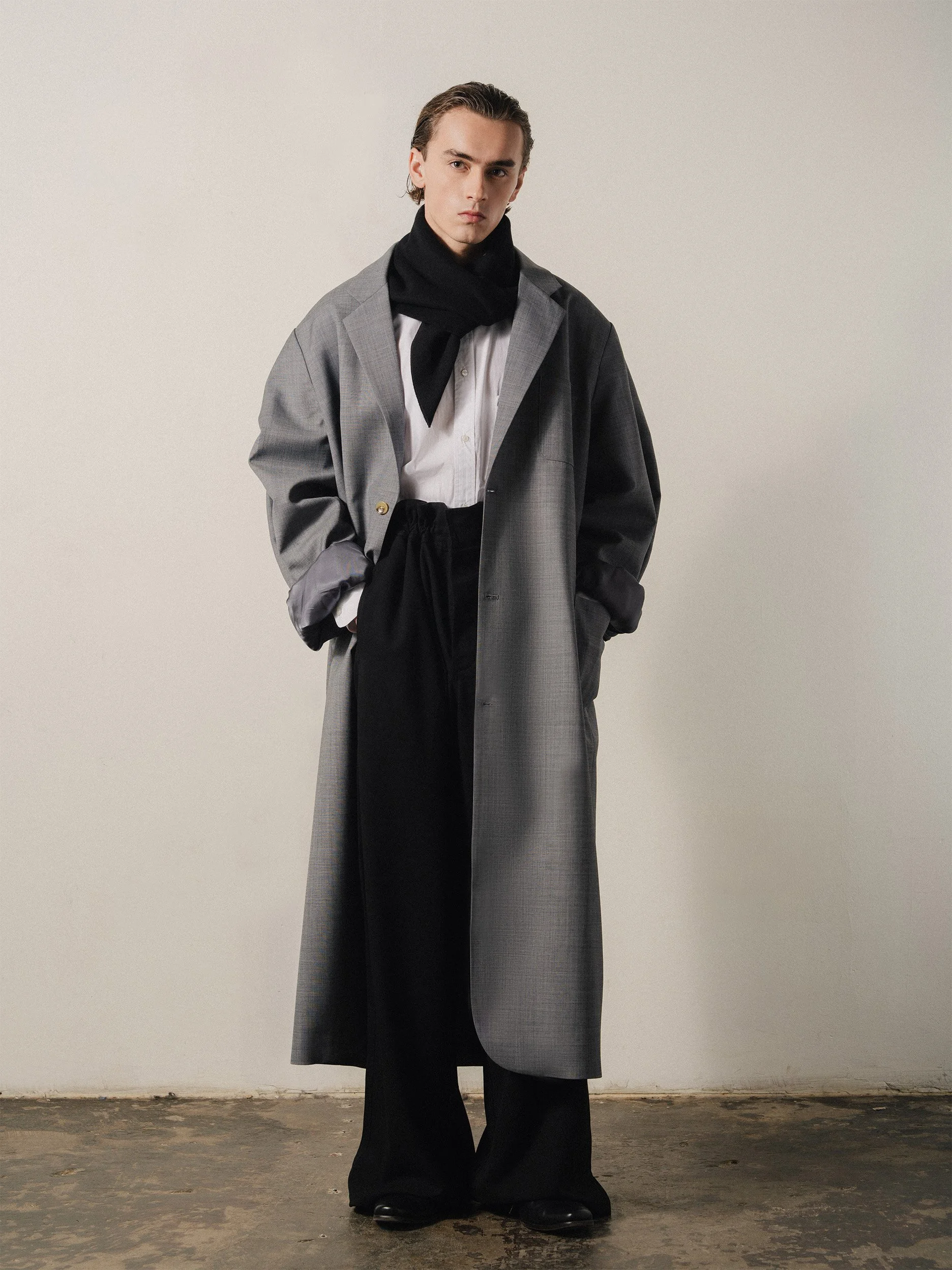 Young man with slicked-back hair wearing a large gray plaid coat over a white shirt, black pants, black shoes, and a black scarf, standing against a plain beige wall.