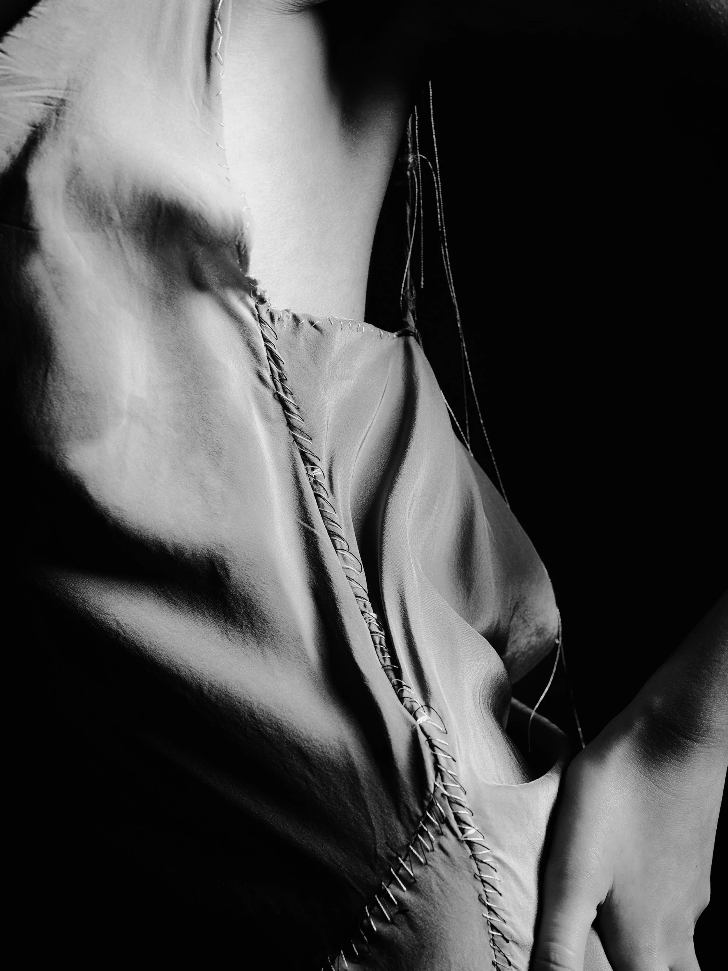 Close-up black and white photo of a person's upper body, showing the shoulder, arm, and part of the chest, with visible stitching on a satin or silk garment.