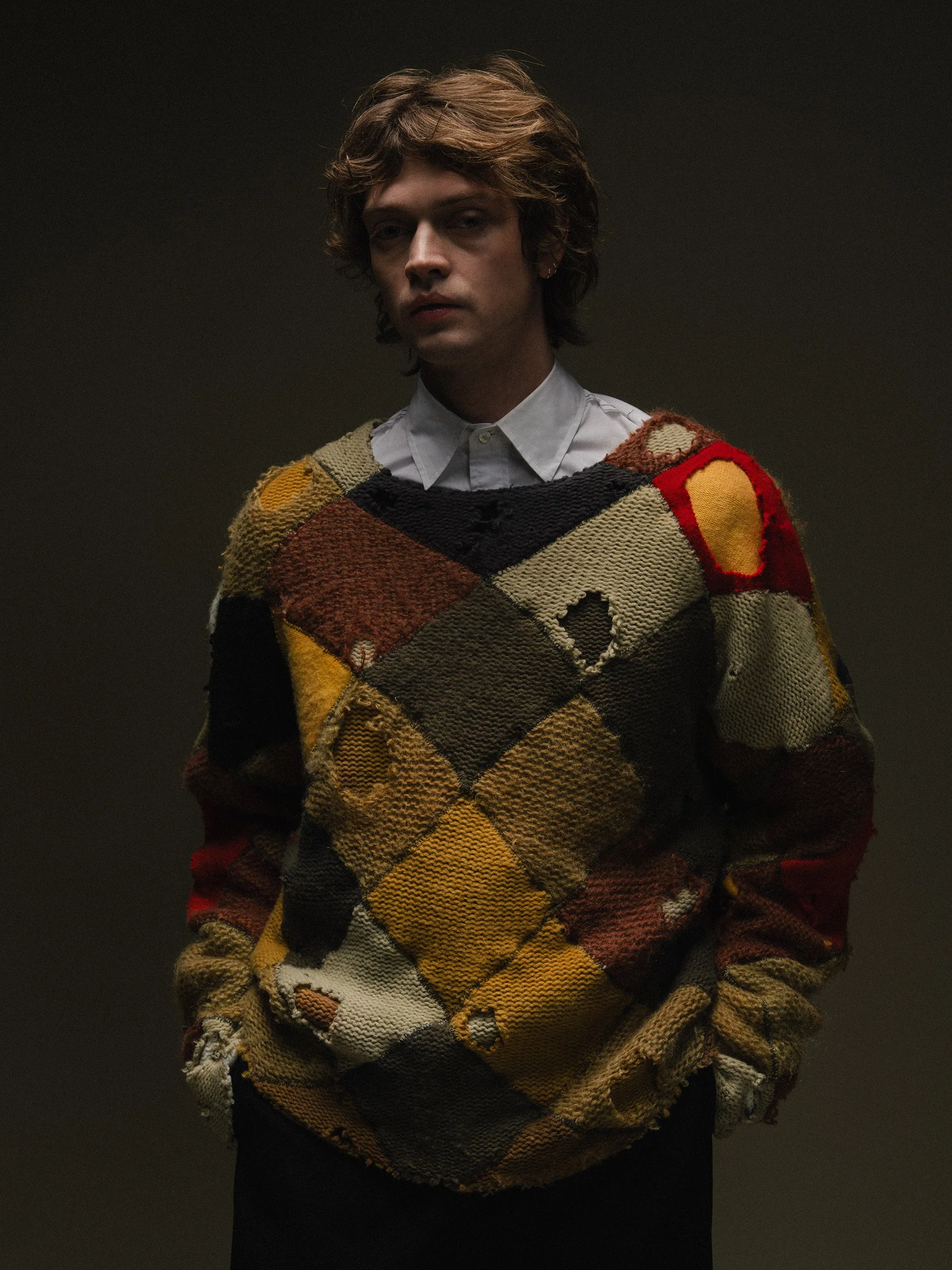 A young man with wavy brown hair wearing a colorful, patchwork knit sweater over a collared white shirt, standing against a plain dark background.