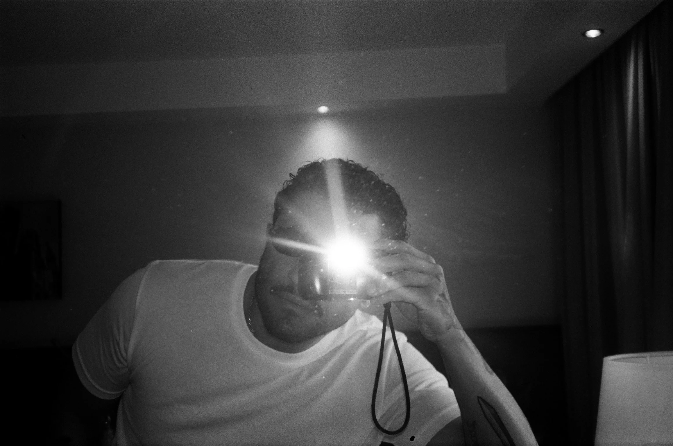 A person taking a selfie in front of a mirror with a bright flash, revealing their face partially obscured by the flash. The person has curly hair and is wearing a light-colored shirt with dark stripes on the sleeves. The room appears to be a dimly lit bedroom or hotel room with curtains on one side and a ceiling with recessed lighting.