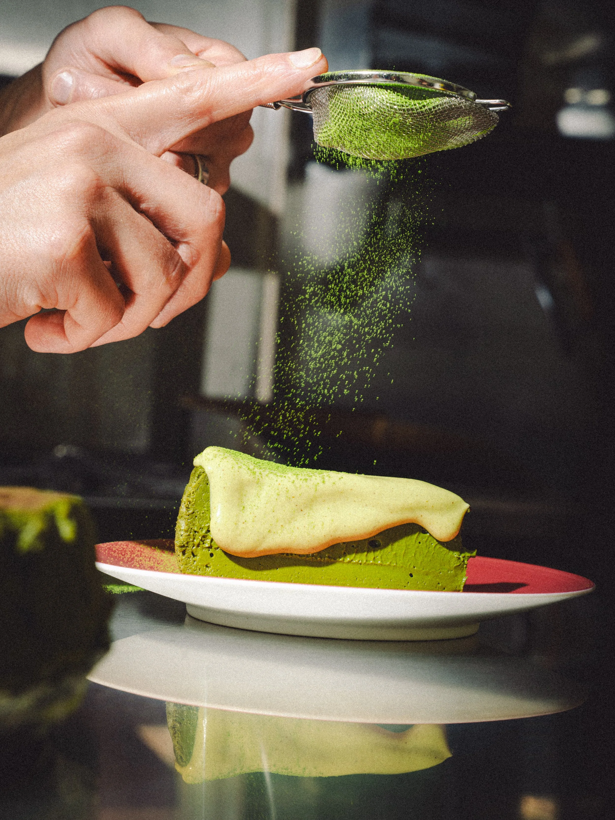 A hand sifting green powder over a slice of green cake on a white plate.