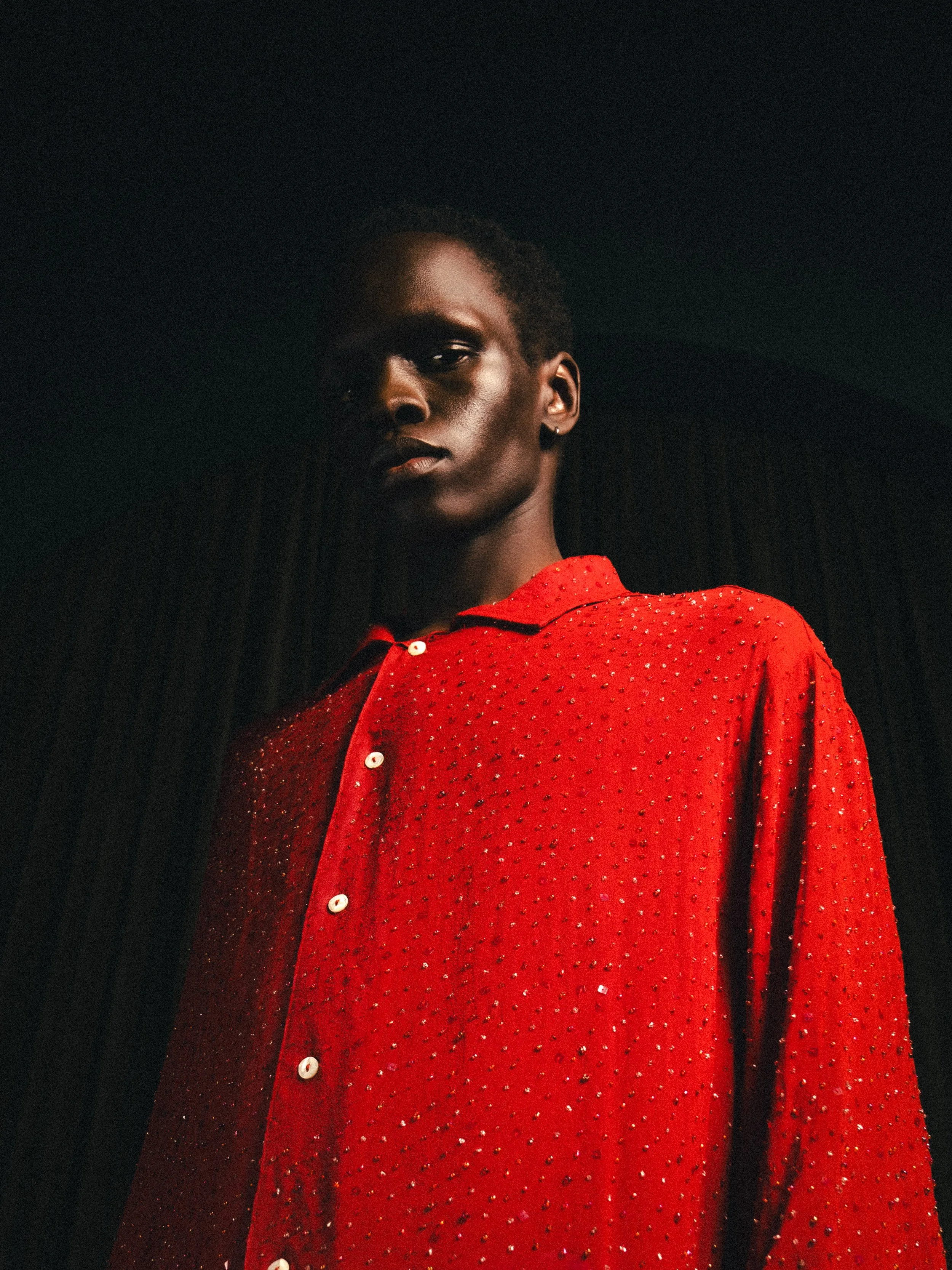 A person wearing a red shirt with sparkling embellishments, buttons, and a collar, standing against a dark background.