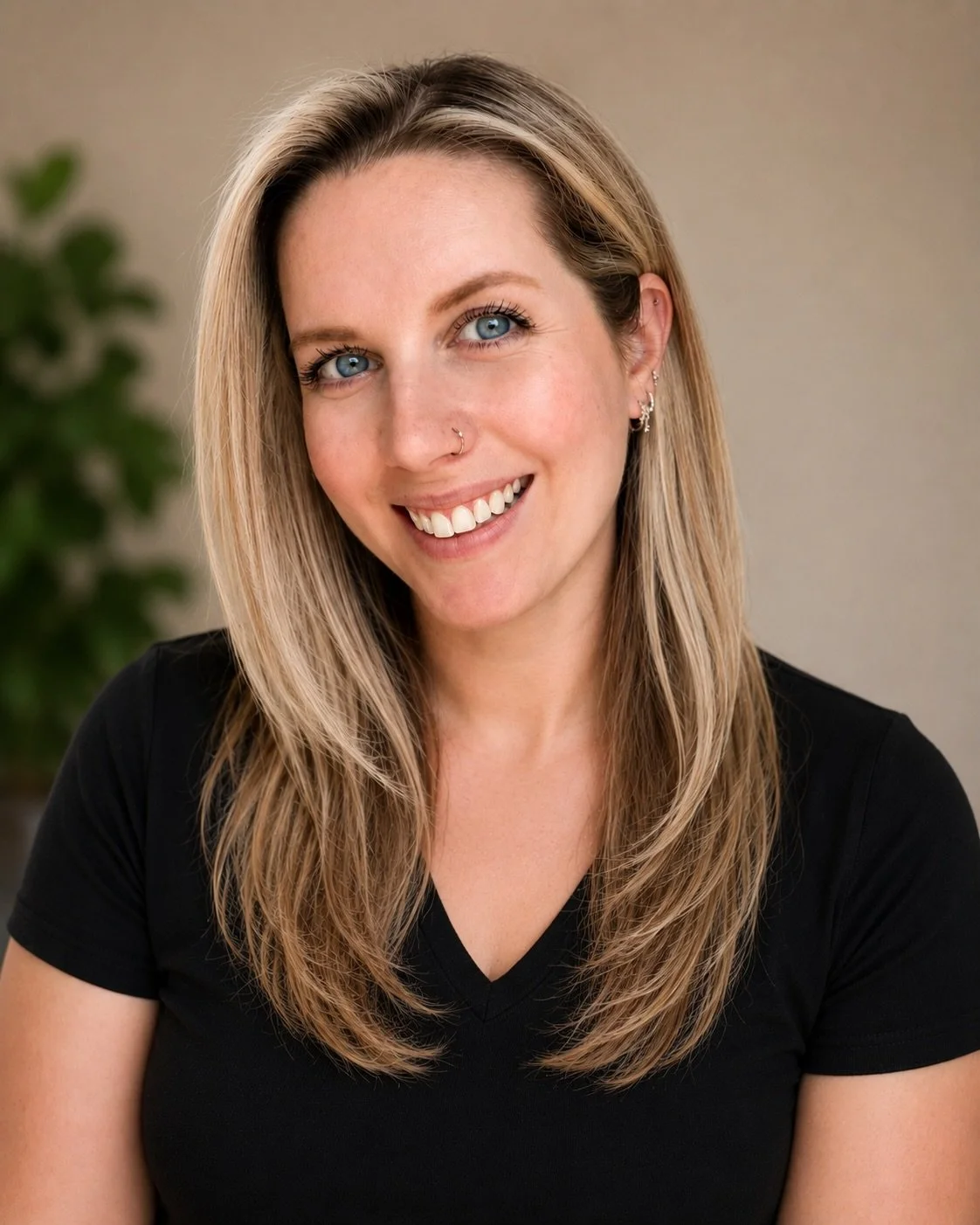 A woman with long blonde hair, blue eyes, and a nose piercings smiling, wearing a black V-neck shirt, with a blurred green plant in the background.