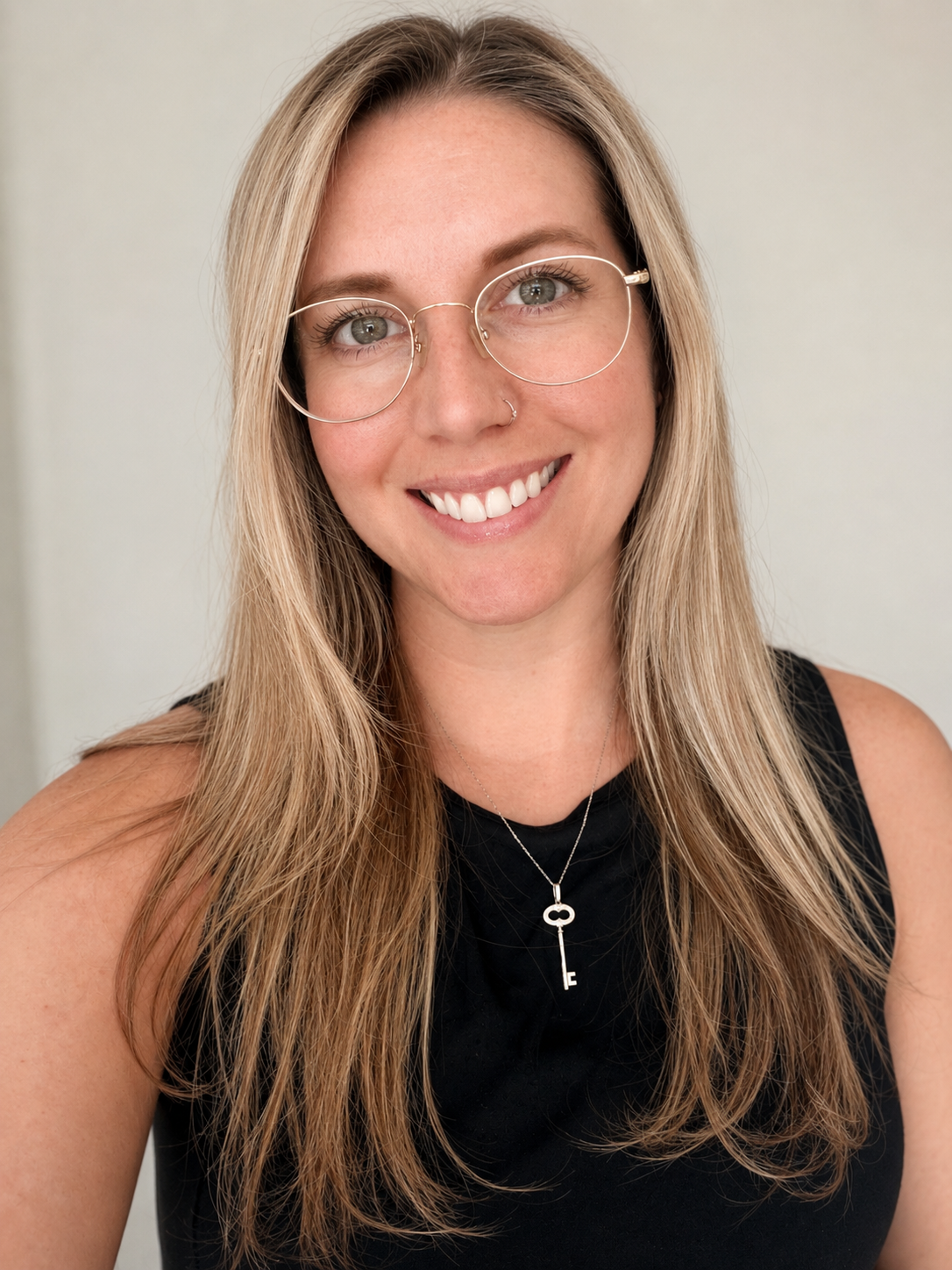 A woman with long blonde hair, glasses, a nose piercing, and a necklace with a key pendant, smiling at the camera against a plain background.