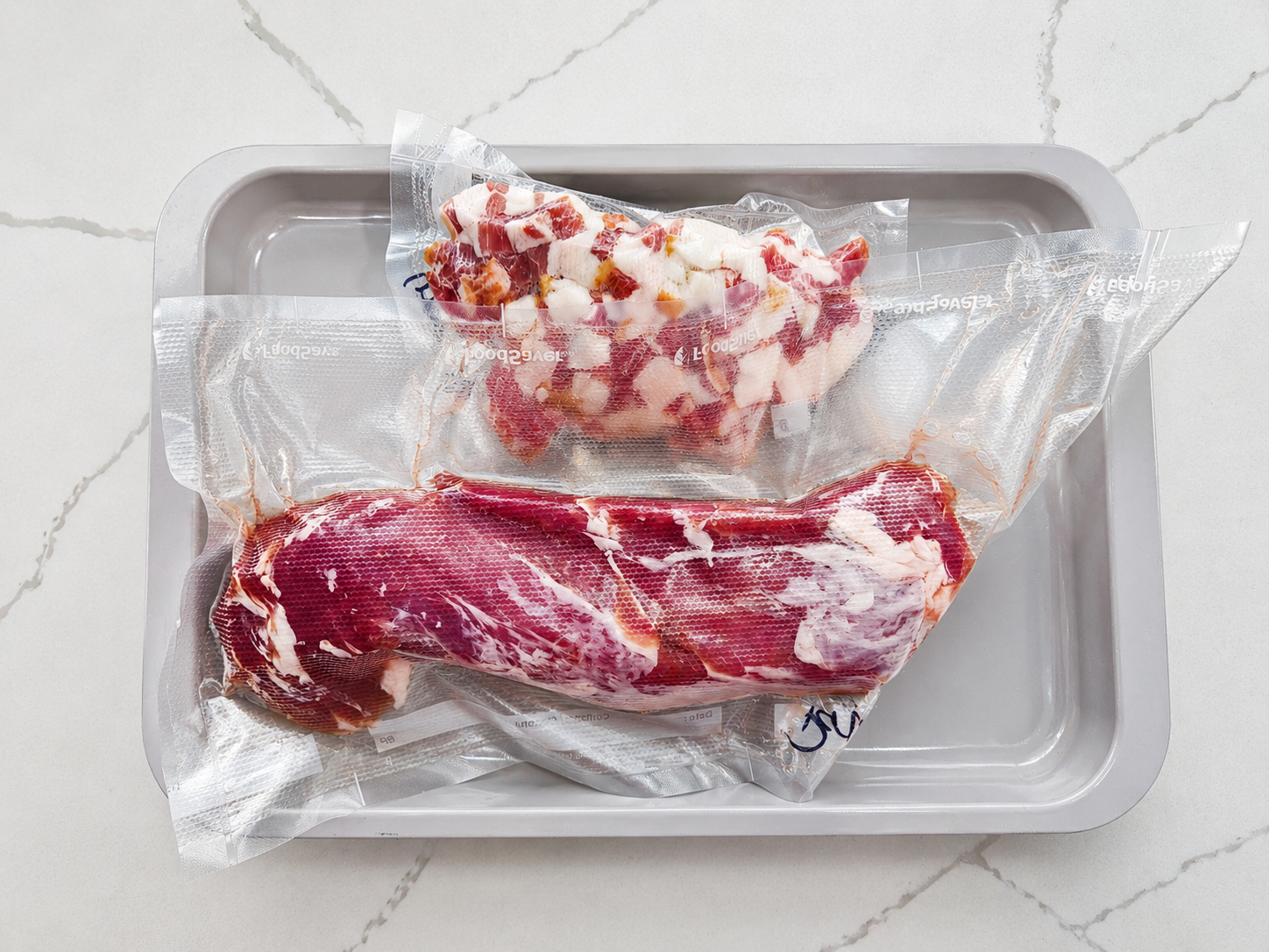 Vacuum-sealed raw meat portions arranged on a rimmed tray over a white marble countertop before thawing.