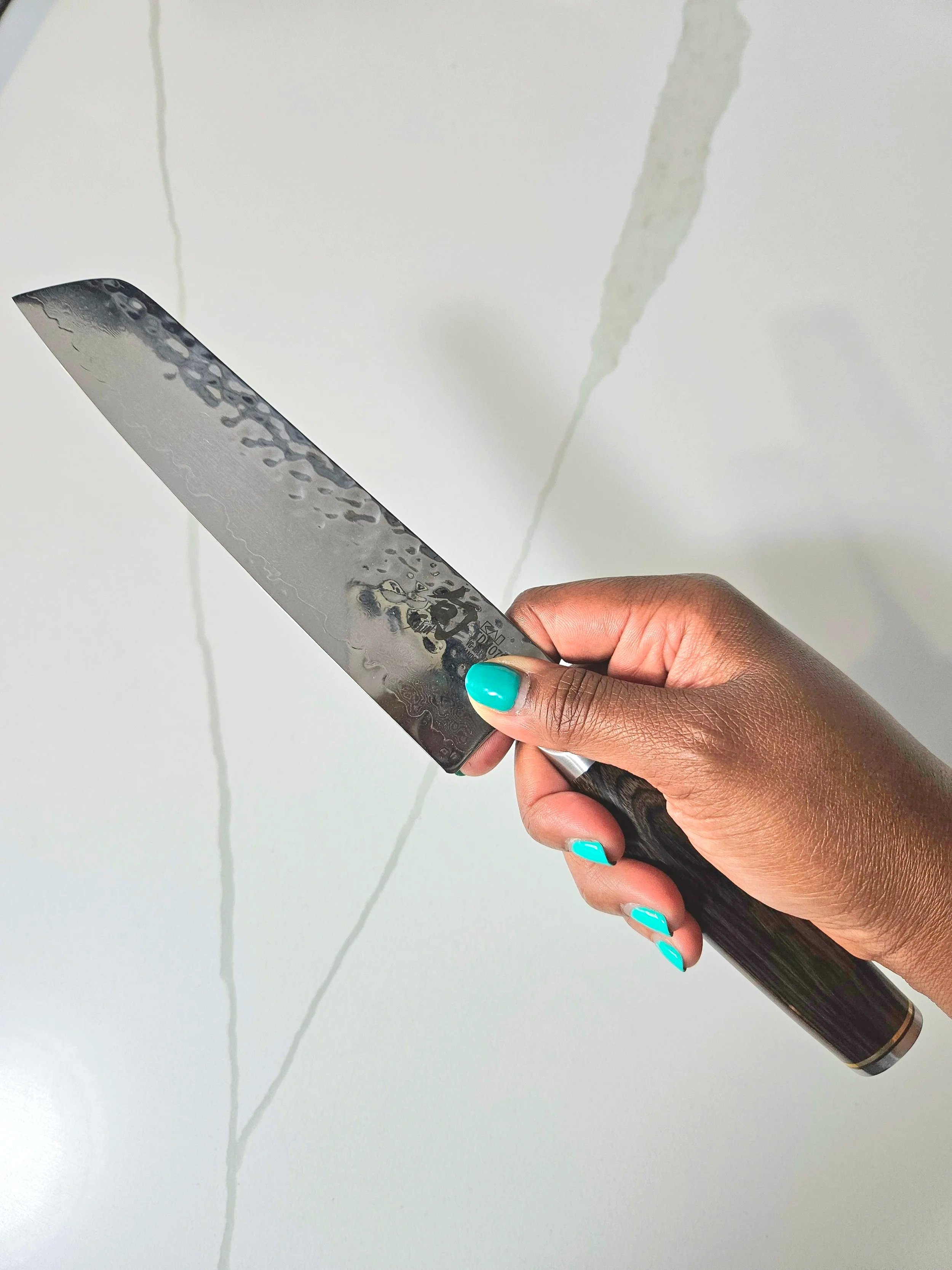 Close-up of a hand with teal nails holding a Close-up of a hand with teal nails holding a Mercer Genesis 9-inch chef’s knife against a bright white marble countertop. against a bright white marble countertop.