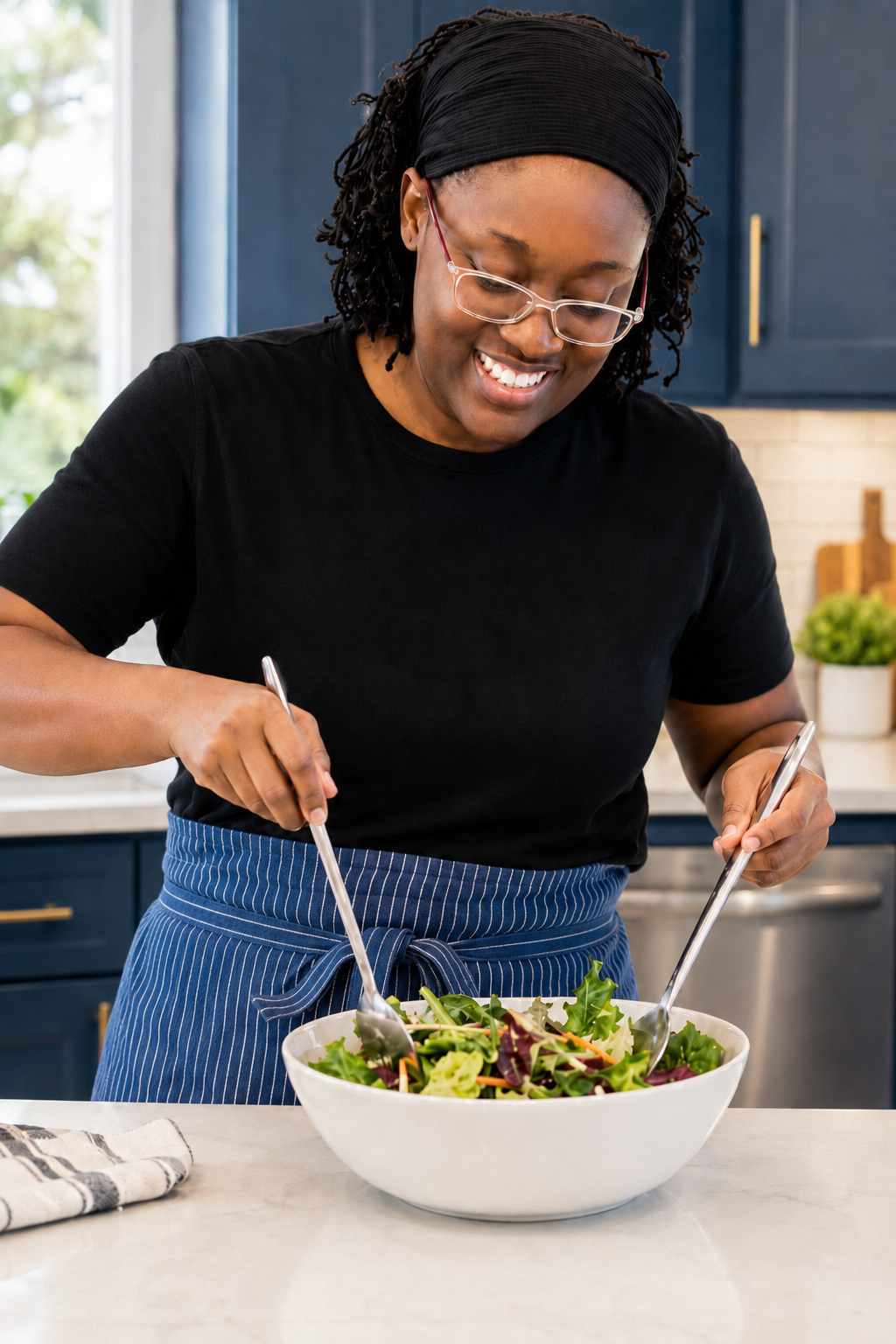 A woman in a black shirt, glasses, and a black headscarf is smiling and tossing a fresh salad in a white bowl in a modern kitchen.
