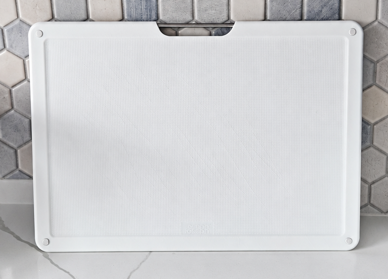 White plastic cutting board displayed upright on a white marble kitchen countertop against a gray and blue hexagon tile backsplash.