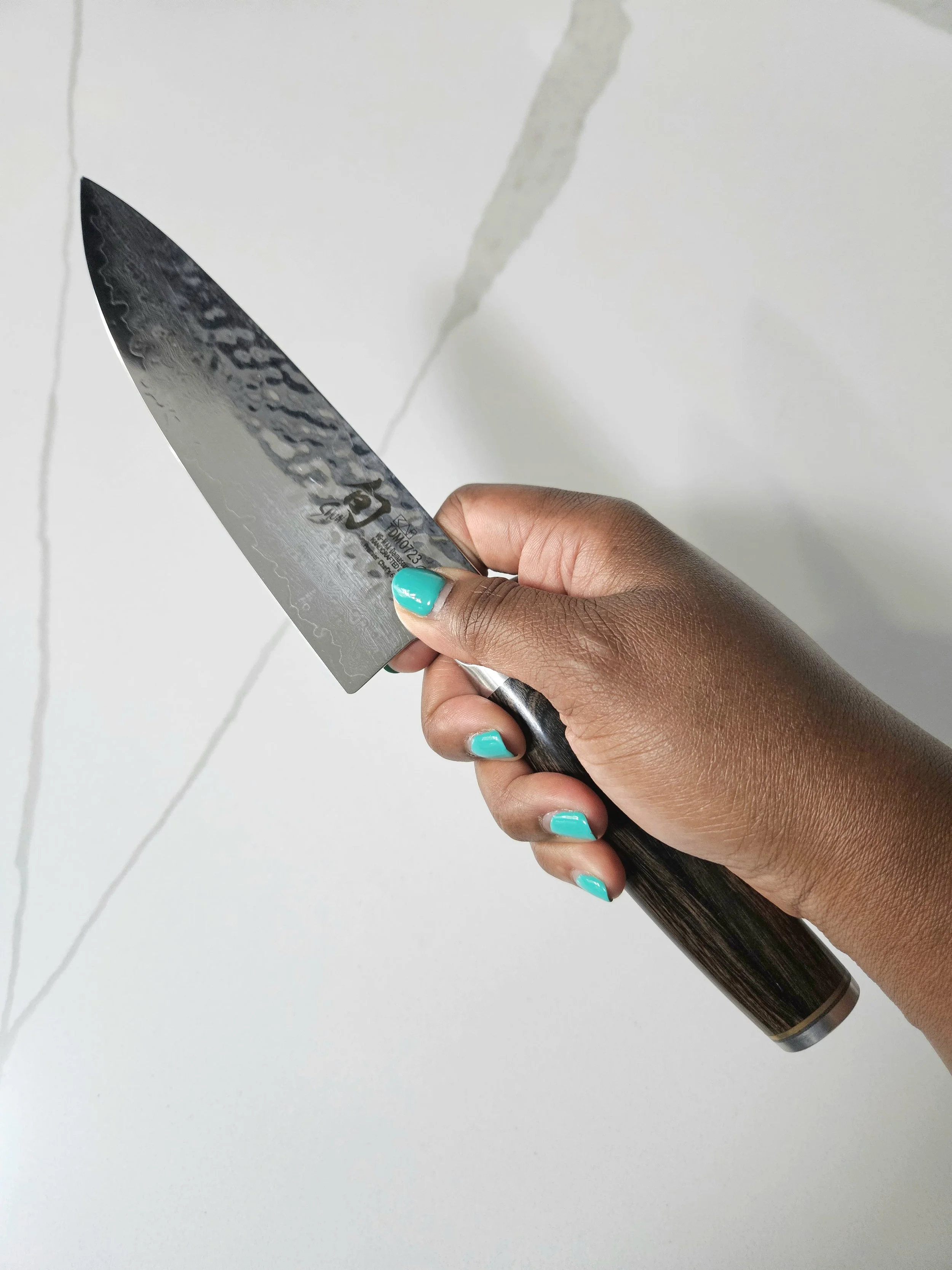 Close-up of a hand with teal nails holding a Shun Premier 6-Inch Chef Knife against a bright white marble countertop.
