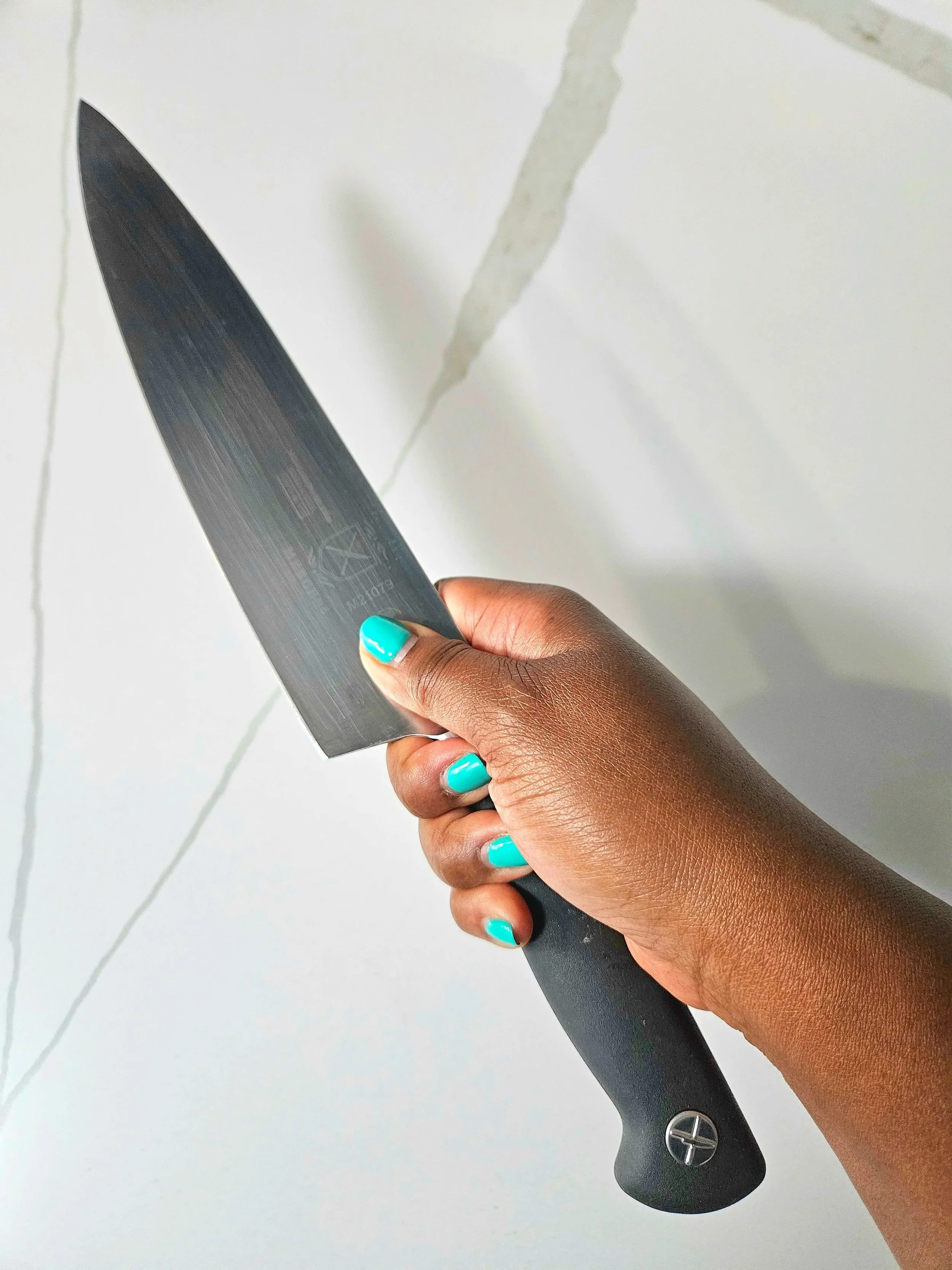 Close-up of a hand with teal nails holding a Mercer Genesis 9-inch chef’s knife against a bright white marble countertop.