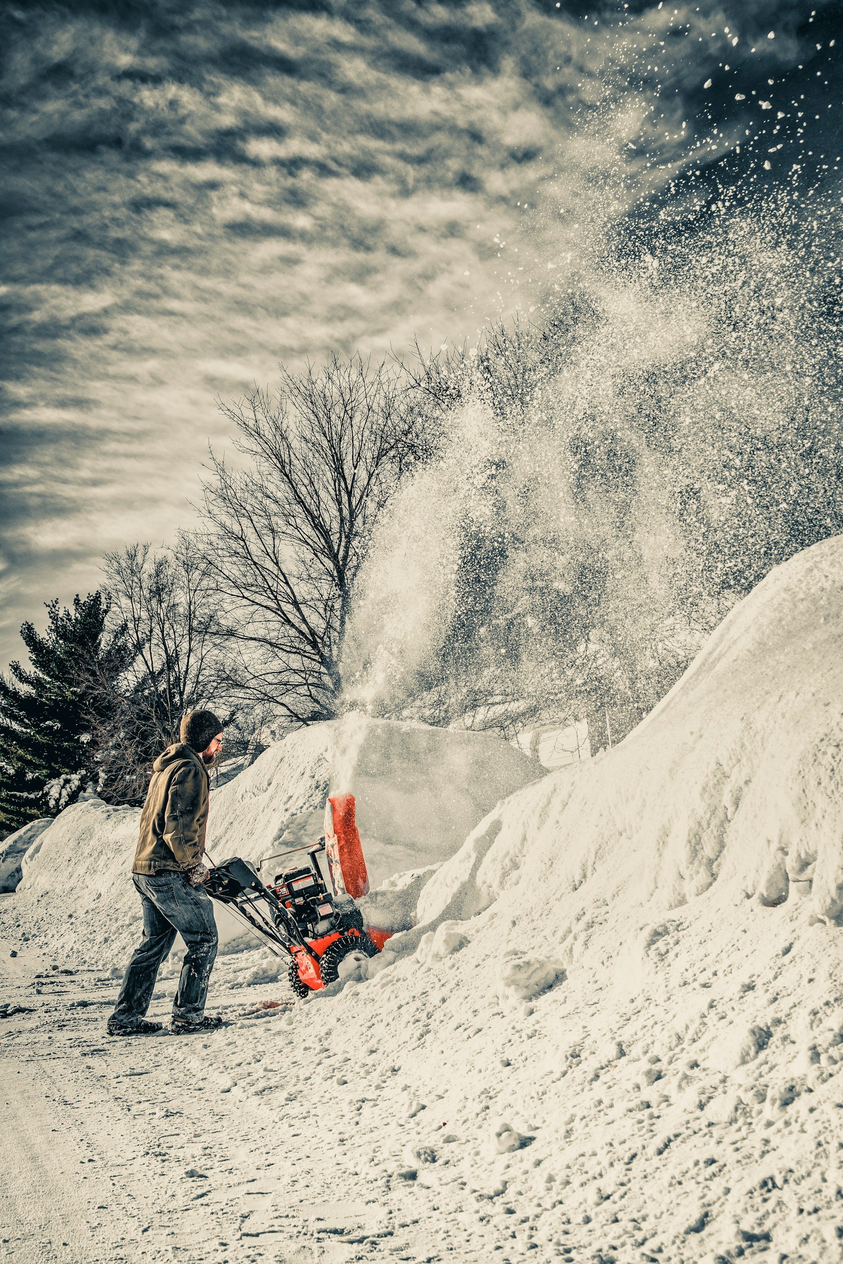 A man in winter clothing and gloves is operating a snowblower on a snowy sidewalk, with large piles of snow in front of him and leafless trees in the background under a cloudy sky.