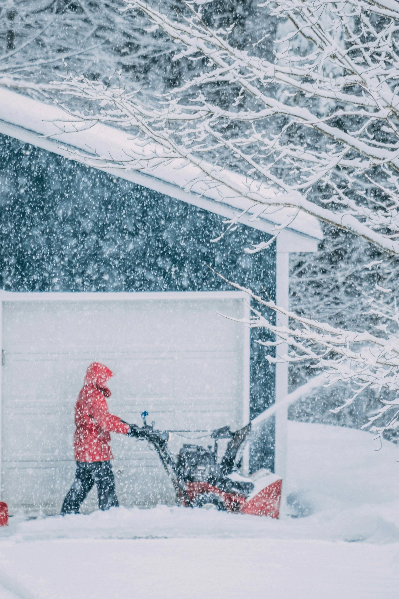 A person dressed in a red coat and dark pants, standing in a snowy yard next to a snow blower, with snow falling heavily and a snow-covered house and trees in the background.