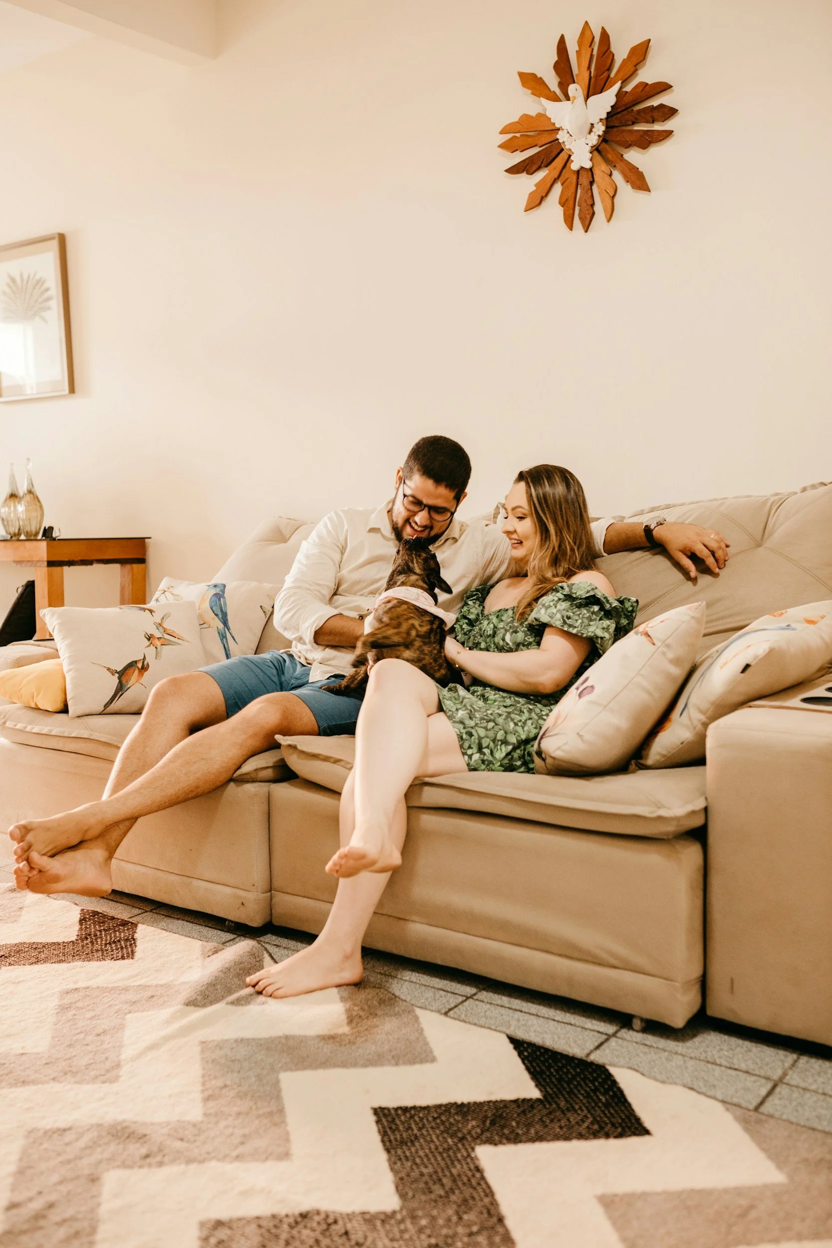 A happy couple sitting on a beige sofa, playing with a dog in a living room. The man is wearing glasses and a white shirt, the woman is in a green dress. Decor includes throw pillows with bird prints, wall art, and a wooden clock on the wall.