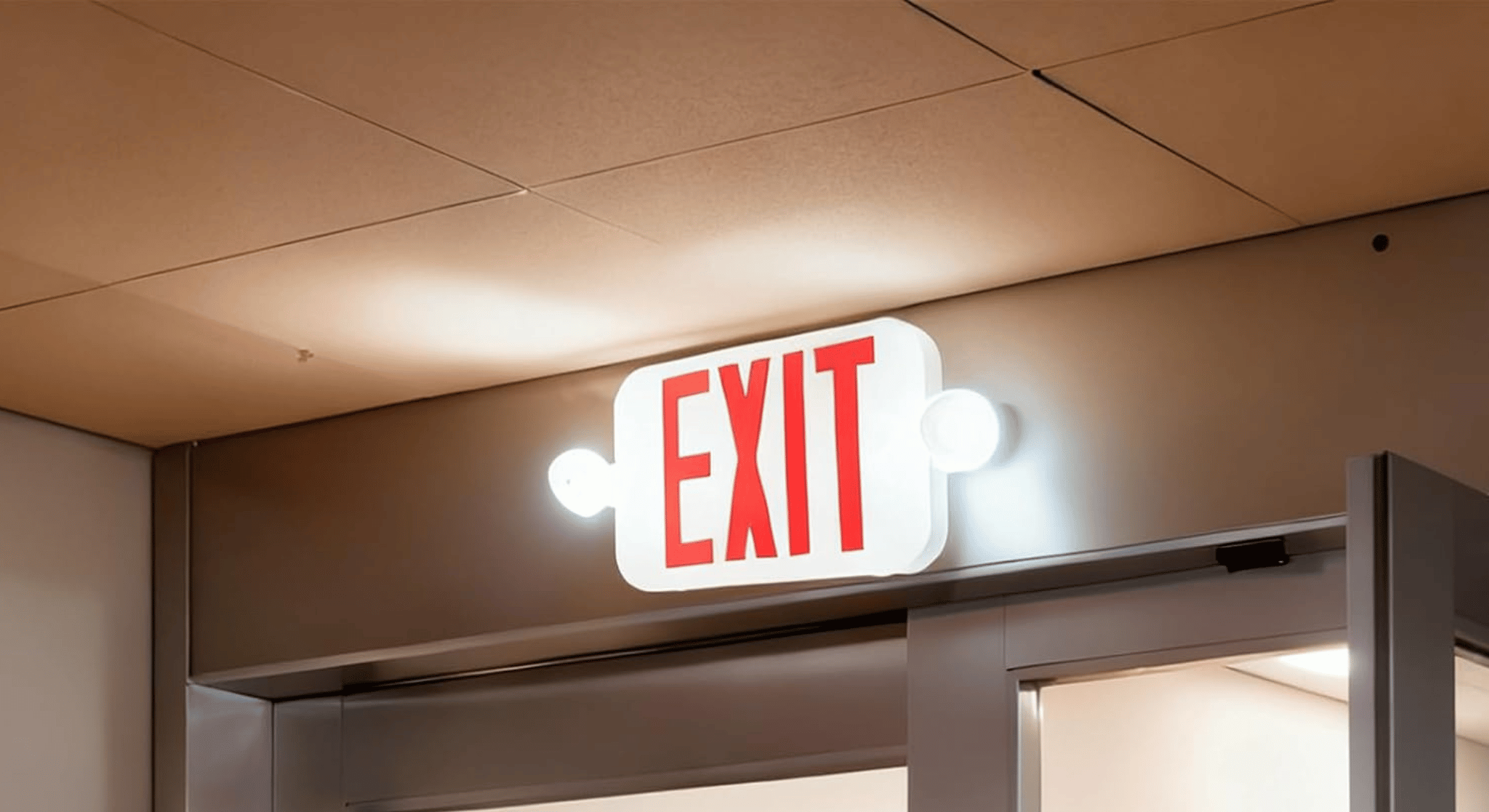 Illuminated white exit sign with red letters above a hallway door in a building.