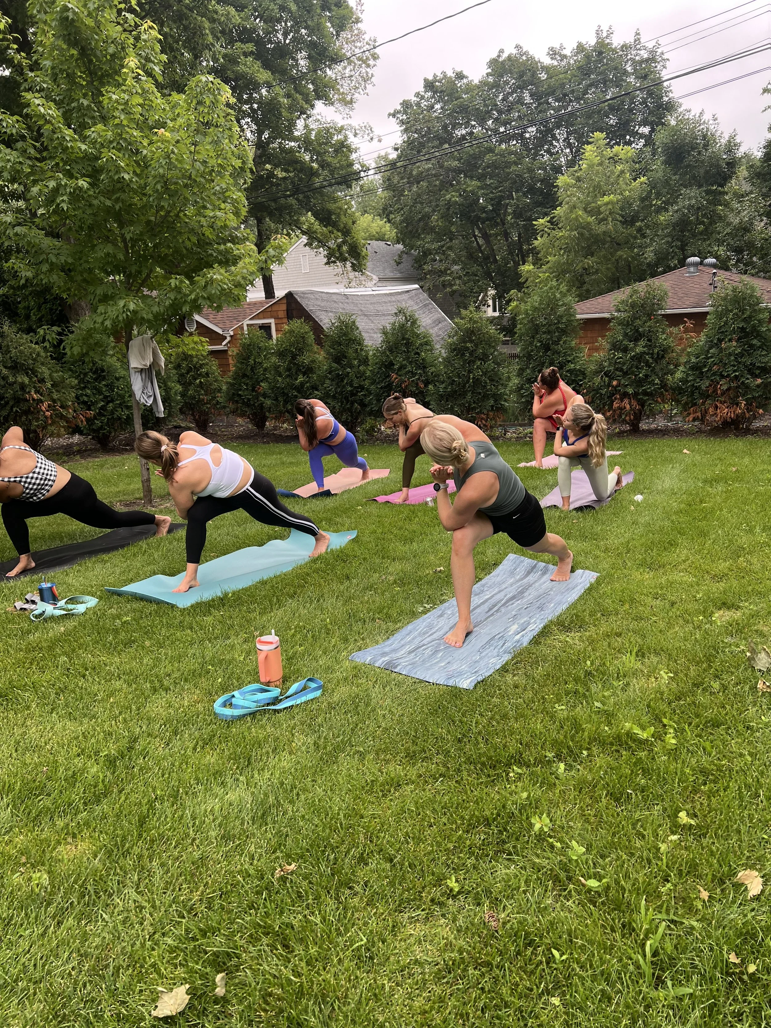 A group of people participating in an outdoor yoga class on a grassy lawn, practicing yoga poses on mats with trees and houses in the background.