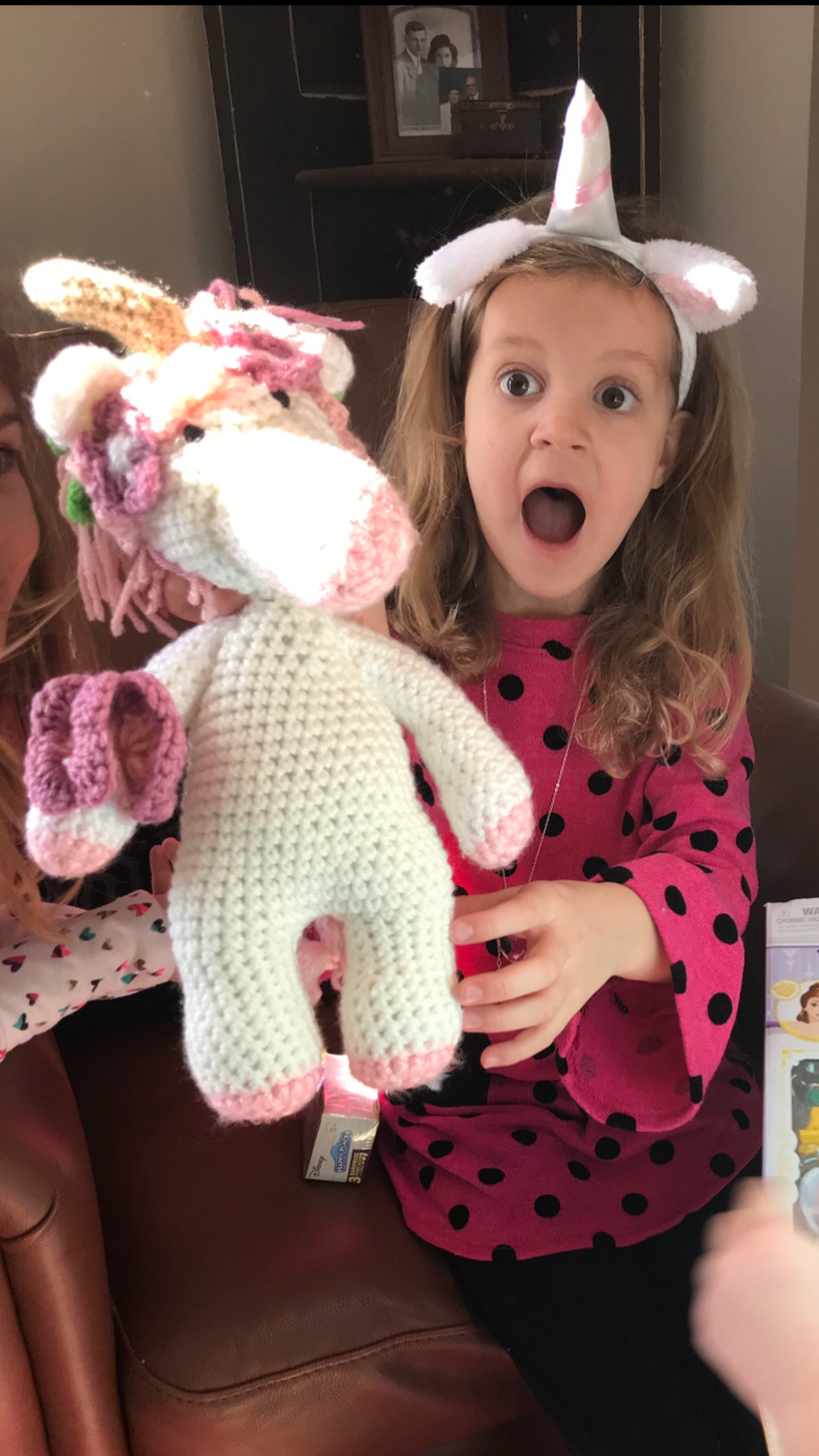 A young girl with surprised expression, wearing a unicorn headband, holding a crocheted unicorn toy, seated on a couch indoors.