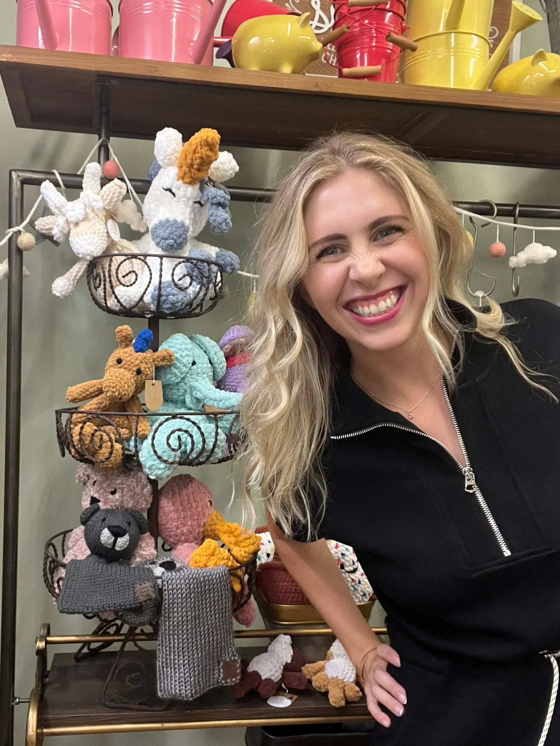 A woman with blonde hair smiling in front of a display of colorful, crocheted stuffed animals and decorative watering cans on a rack.