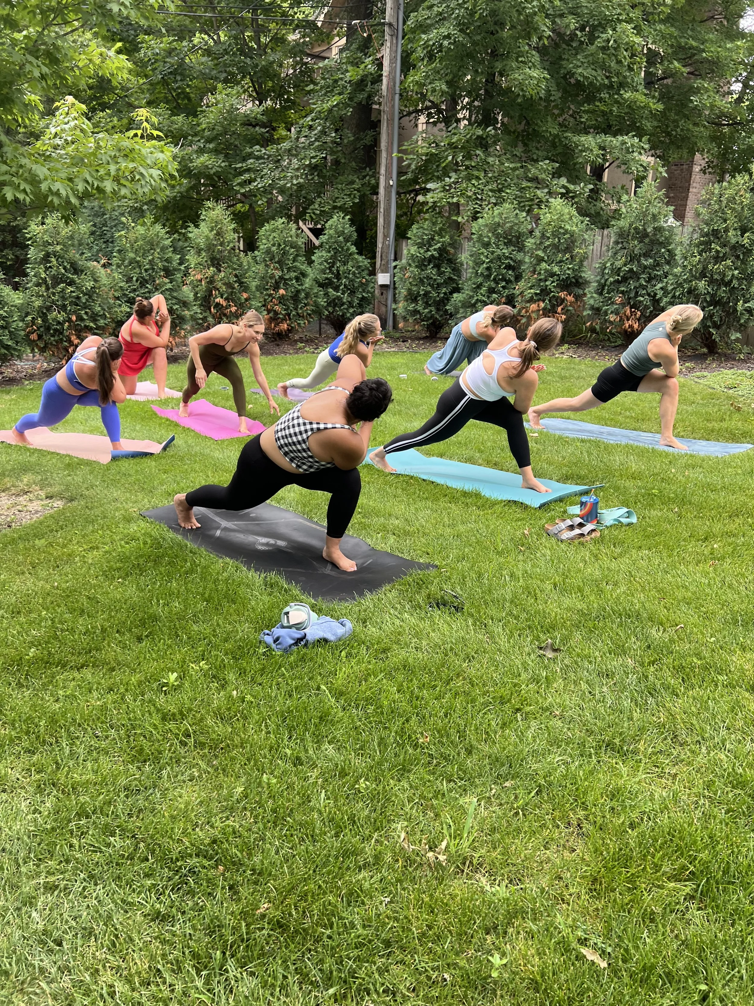 A group of women practicing yoga outdoors on yoga mats on a grassy lawn, with trees and bushes in the background.