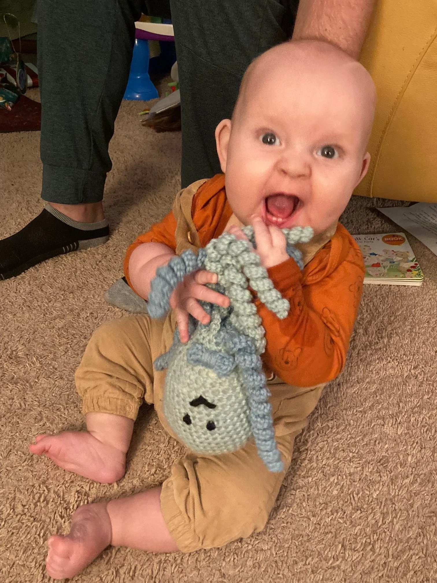 A young child with a bald head and wide eyes, sitting on a carpeted floor and holding a knitted gray and white stuffed animal with a snowman face. An adult's hand is touching the child's foot.