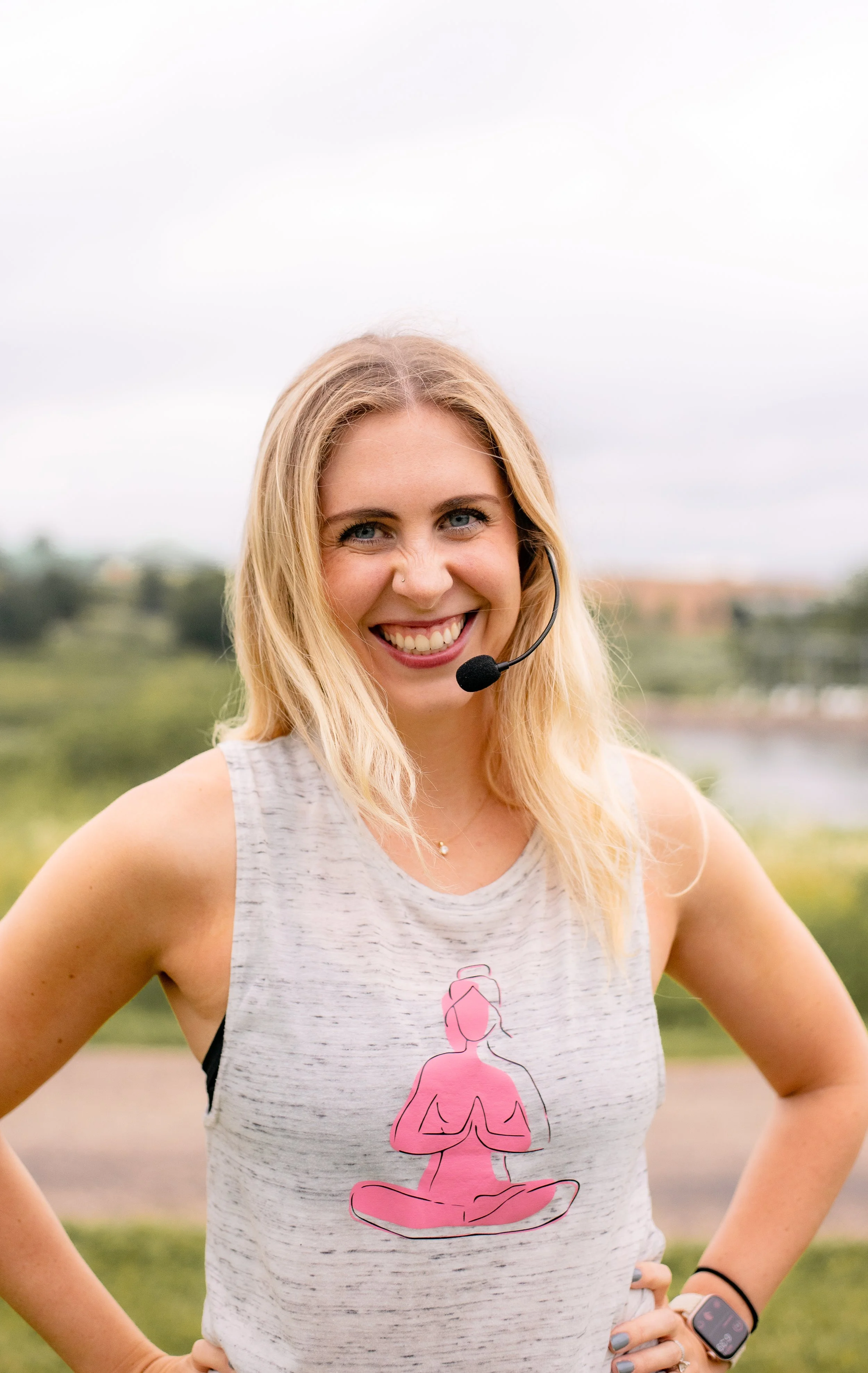 A young woman with blonde hair and blue eyes smiling outdoors, wearing a gray tank top with a pink yoga figure design, a smartwatch, and a headset against a cloudy sky and green landscape.