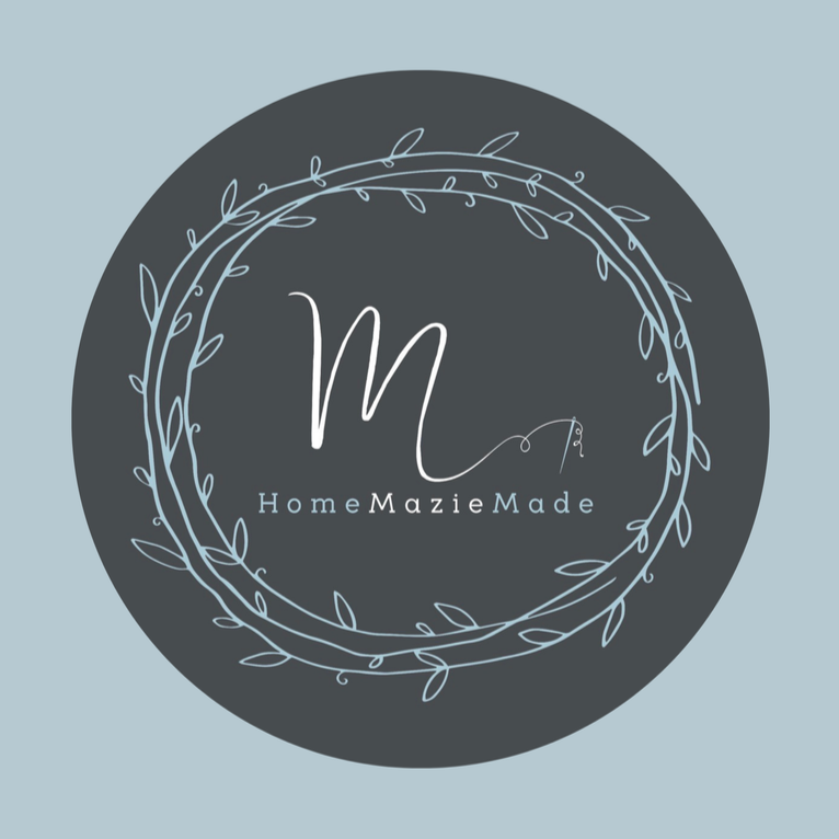 A circular logo with a dark gray background and a light blue, leaf-wrapped border. In the center, there is a stylized letter 'M' and the text 'Home Mazié Made' underneath.
