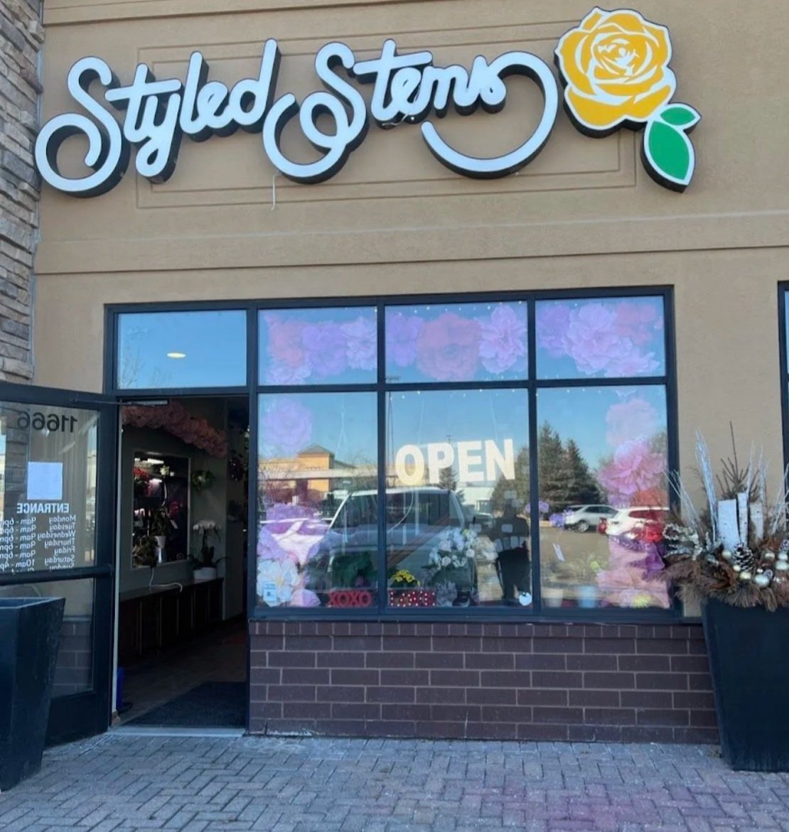 Exterior of a flower shop named 'Styled Stems' with a large window displaying an 'OPEN' sign, and decorated with flowers and plants.