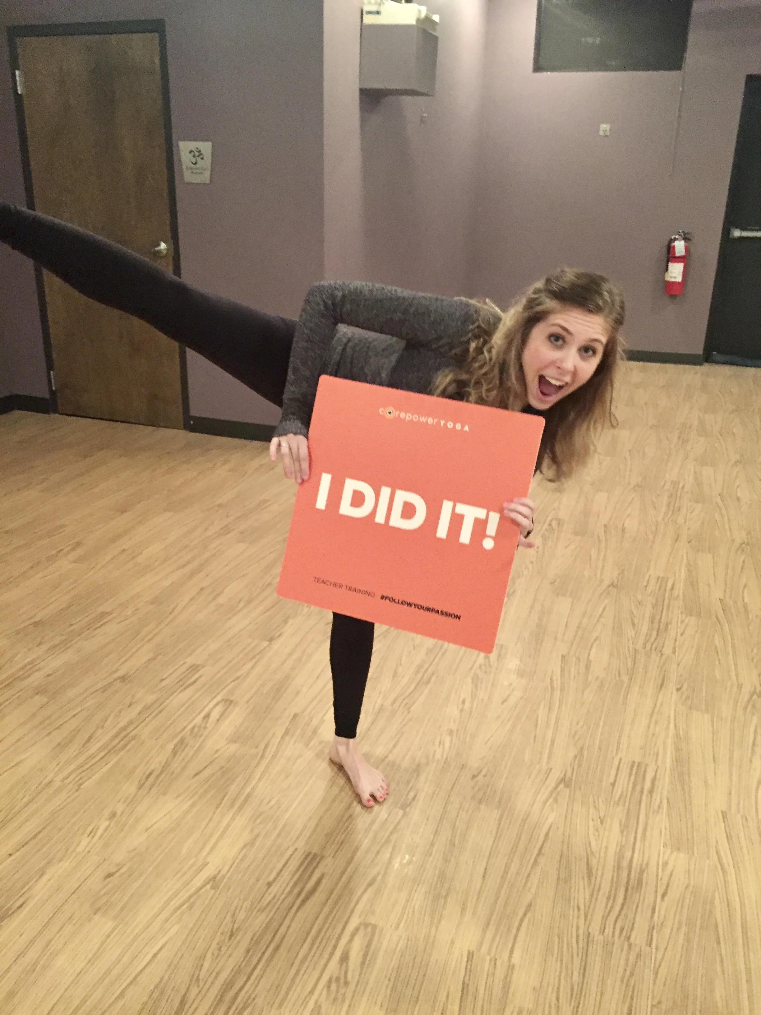 A woman is performing a move in yoga studio, balancing on one foot with her other leg extended horizontally, holding an orange sign that reads 'I DID IT!'. She appears excited and happy, with an open-mouthed smile,. She is barefoot, wearing dark leggings and a gray shirt.