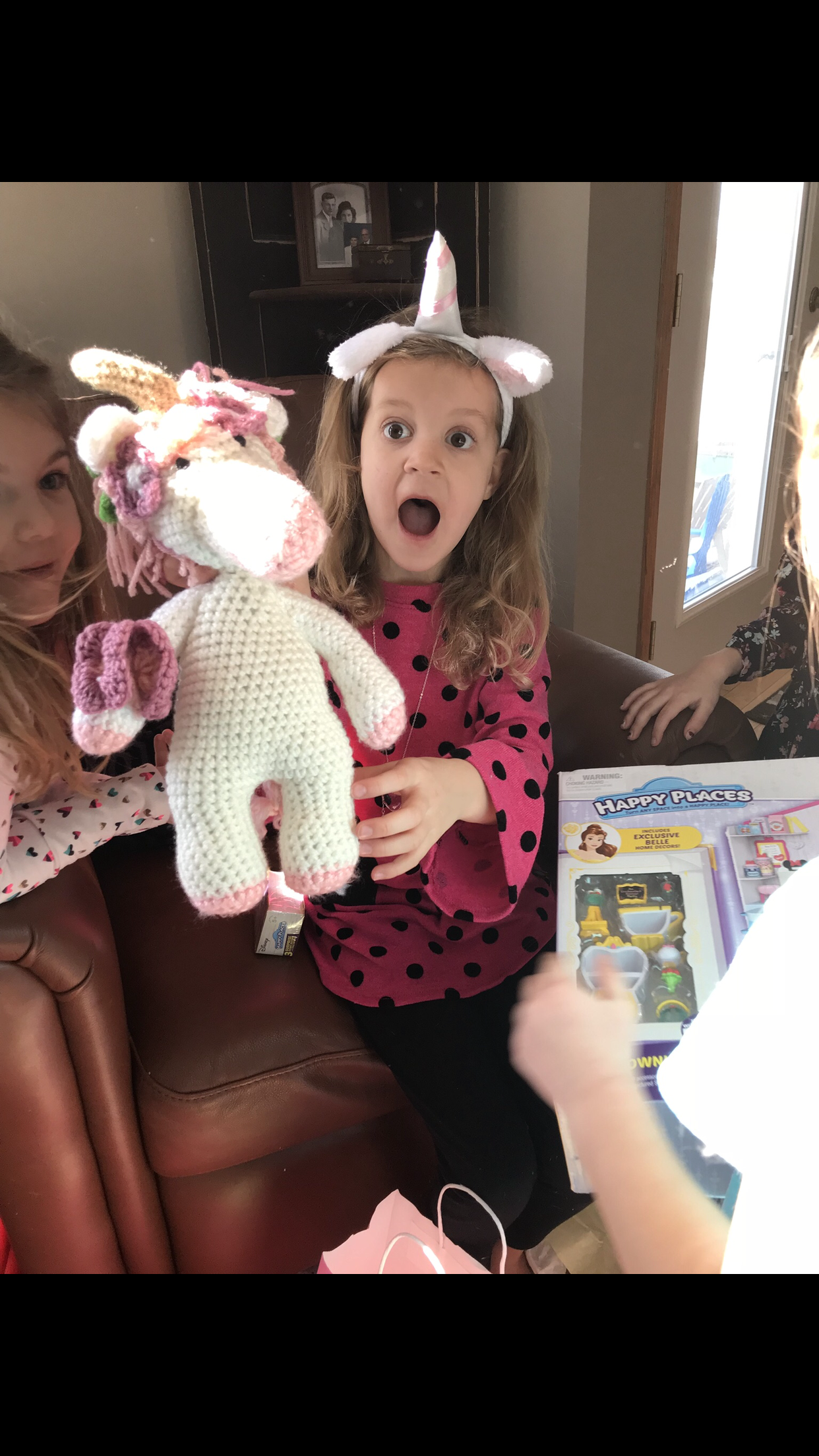 A young girl wearing a unicorn headband with a surprised expression on her face, holding a crochet unicorn stuffed toy. Other children and toys are visible around her in a room with a window and family photos.
