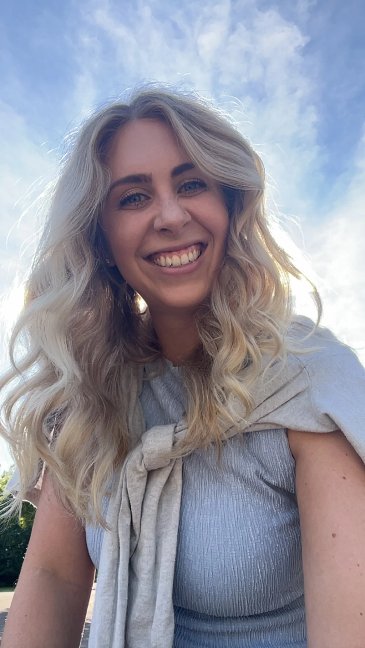A smiling woman with long, wavy blonde hair and blue eyes, outdoors with a partly cloudy sky in the background.