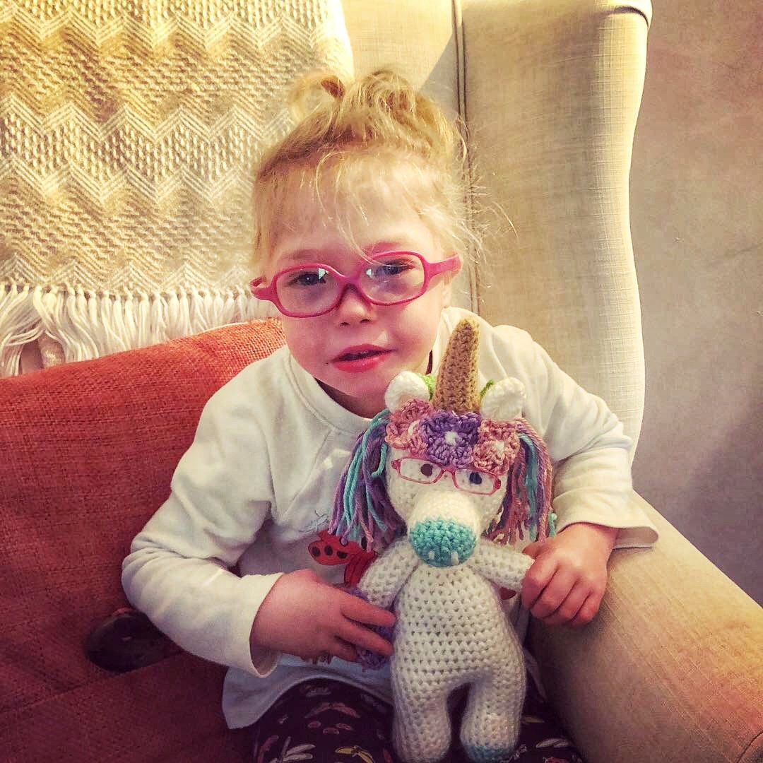 A young girl with red hair, pink glasses, and a white shirt sitting on an orange couch, holding a colorful crocheted unicorn stuffed animal.