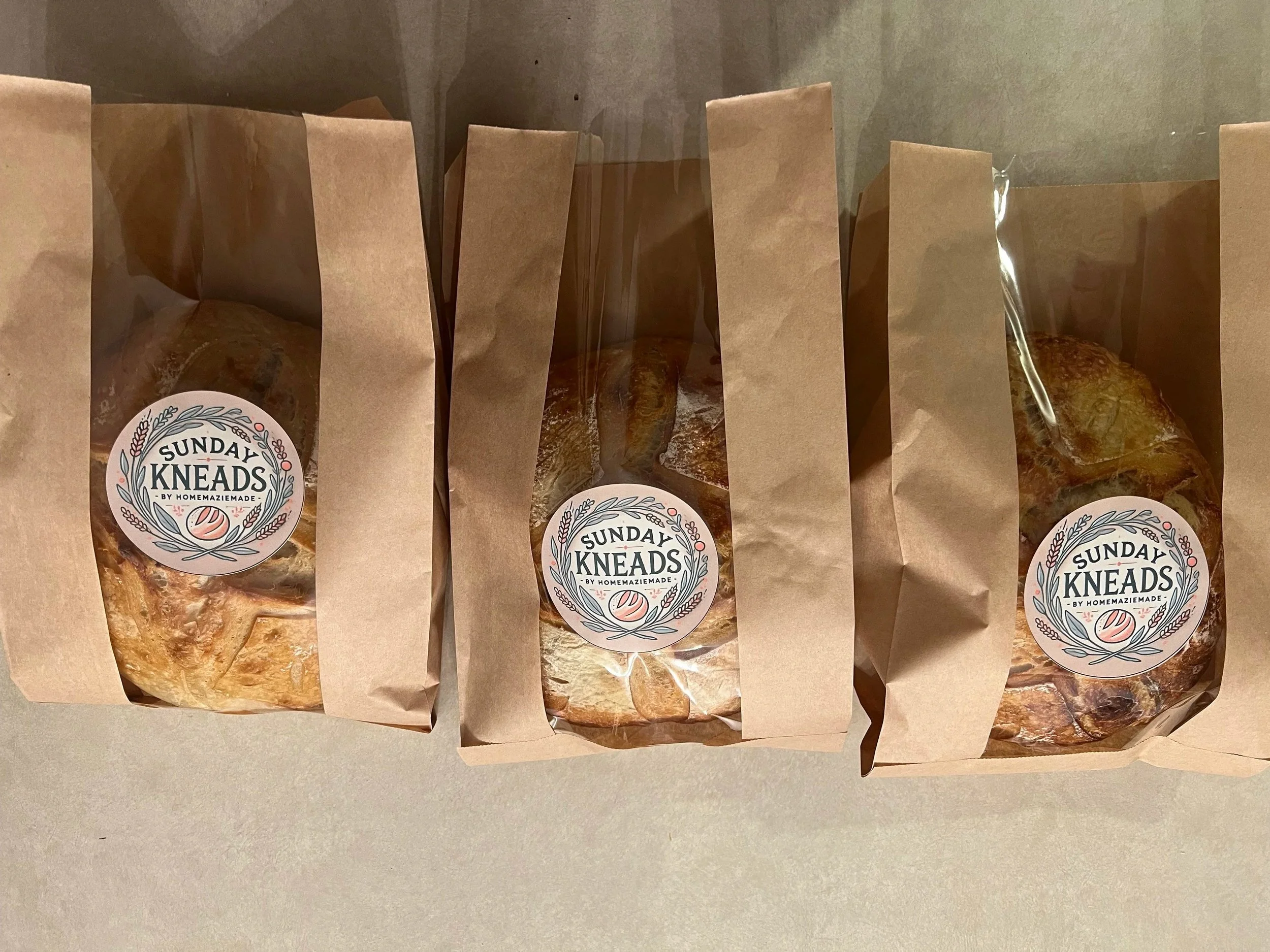 Multiple brown paper bags labeled 'Sunday Kneads by HomemazieMade' containing baked goods, lined up on a gray surface.