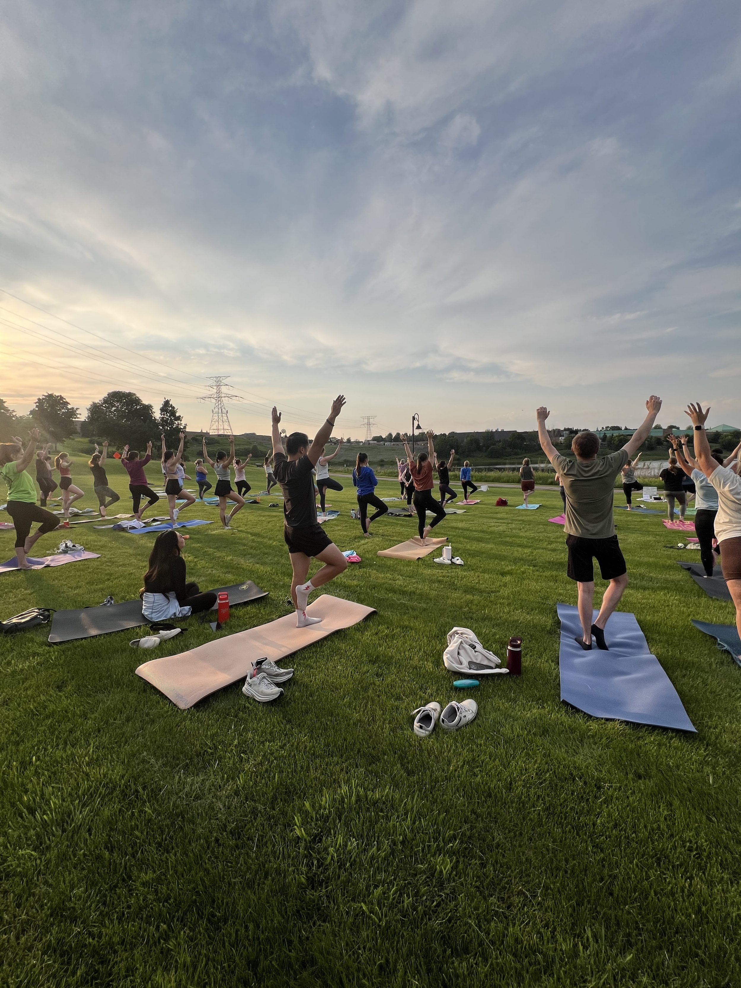 Group of people participating in outdoor yoga class on a grassy field during sunset, with power lines and a few trees in the background.