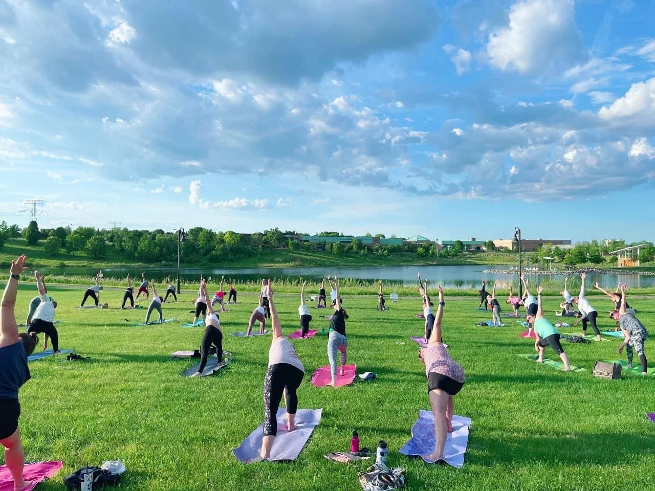 Group of people practicing yoga outdoors on a grassy field near a lake, with trees and buildings in the background and a partly cloudy sky overhead.